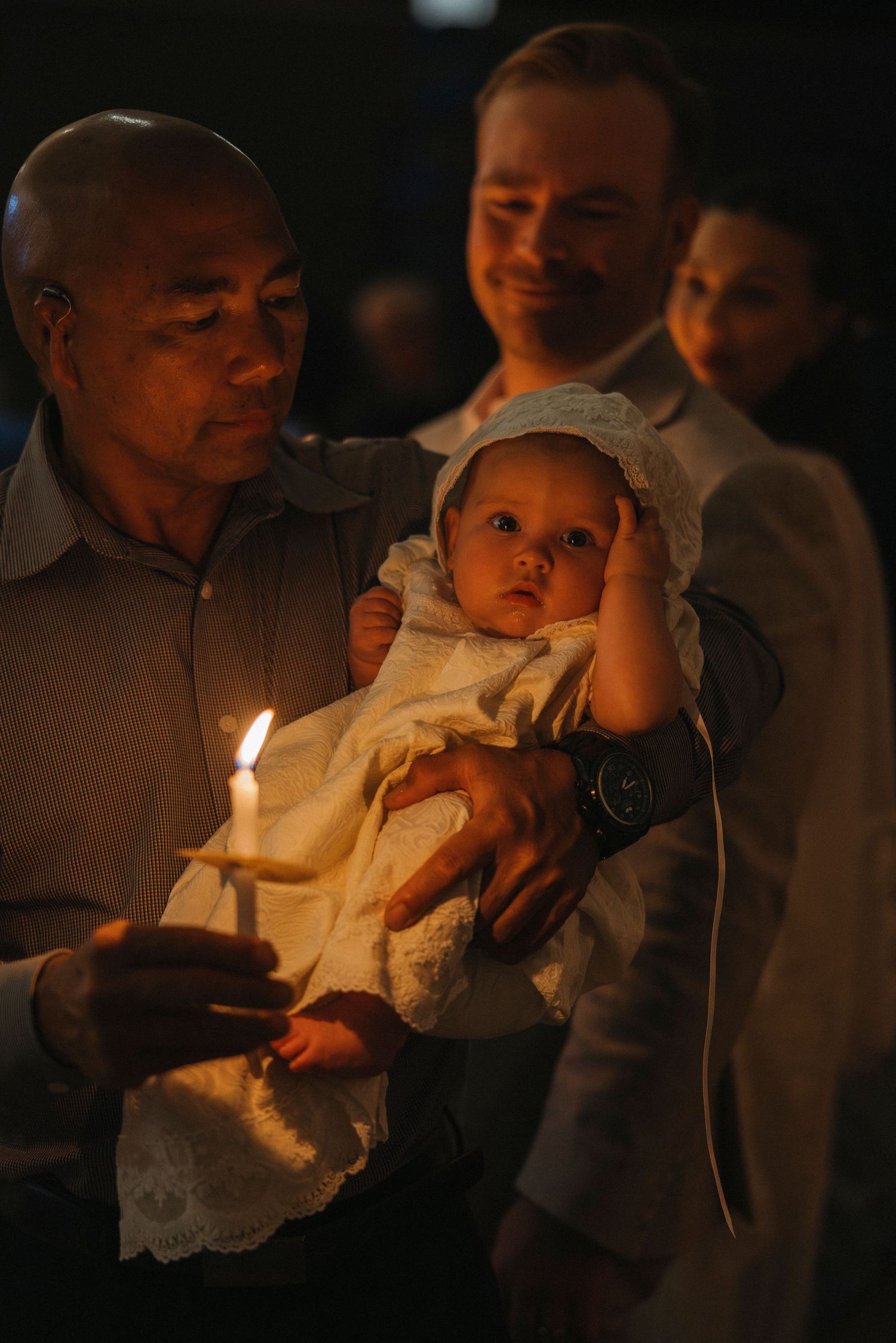 Man holding baby in baptismal gown with lit candle during Catholic baptism ceremony.
