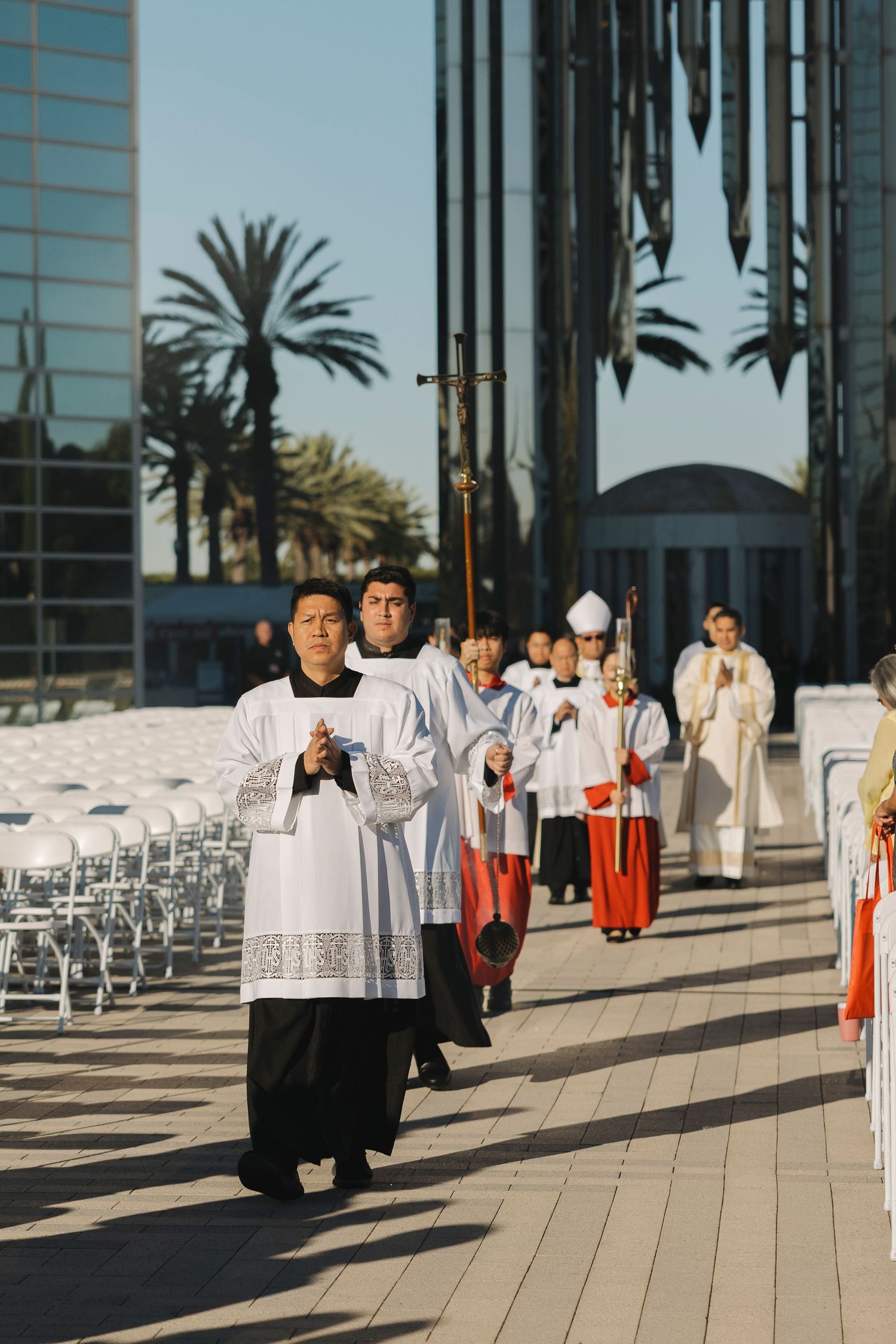 Religious procession with altar servers in white robes and red garments walking outside a glass building.