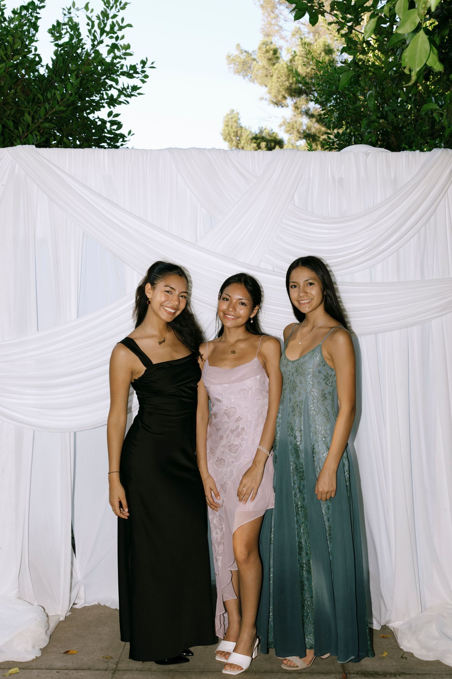 Three women in formal dresses pose against a white draped backdrop at an outdoor event.