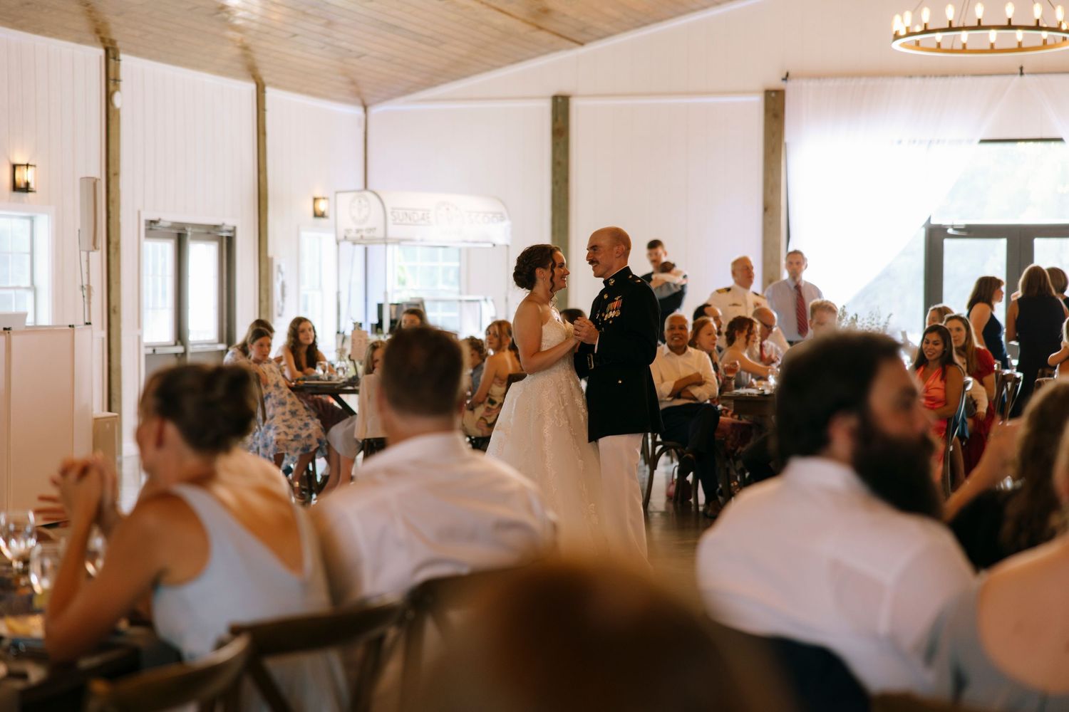 Bride and groom share their first dance at a rustic wedding reception while guests watch from decorated tables.