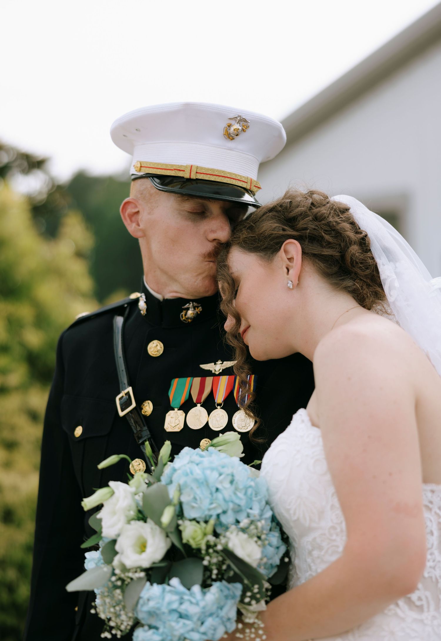 Marine officer in dress uniform kisses bride on forehead, she holds blue hydrangea bouquet at outdoor wedding ceremony.
