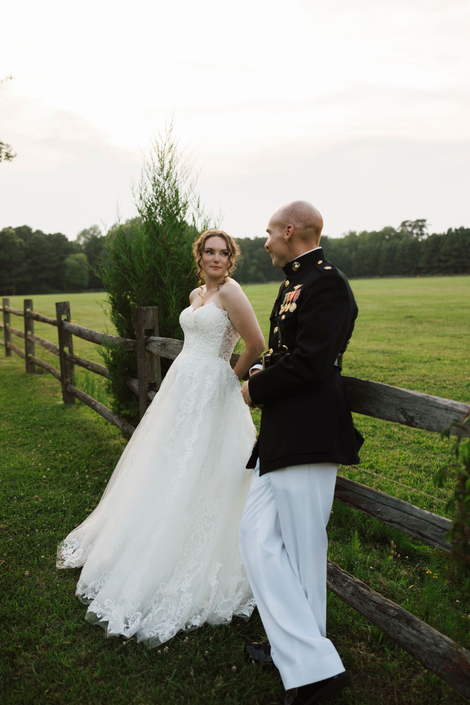 Military groom in dress uniform holds hands with bride in white lace gown by rustic fence at outdoor wedding.
