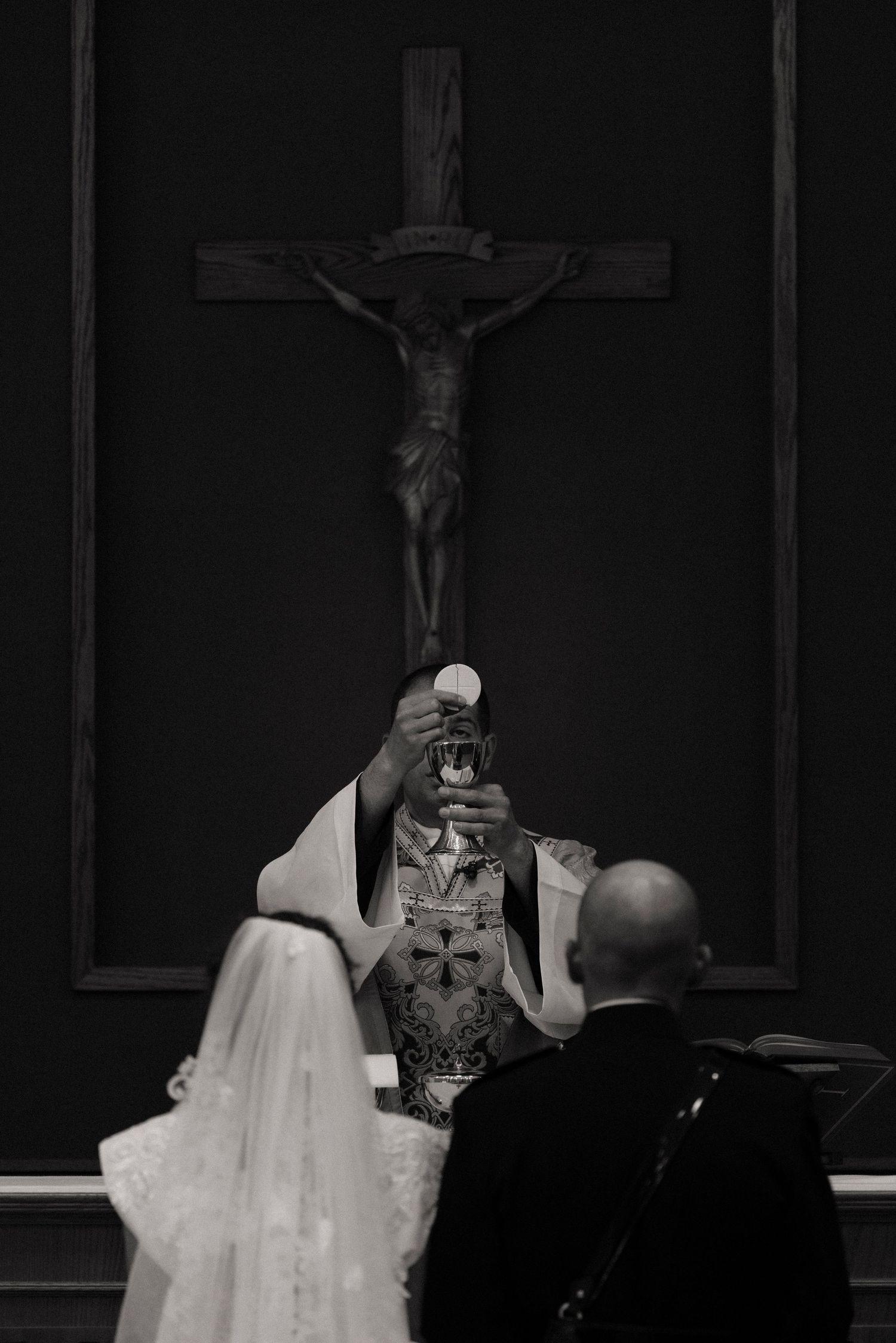 Black and white photo of a Catholic wedding ceremony with priest elevating the Eucharist before a crucifix.