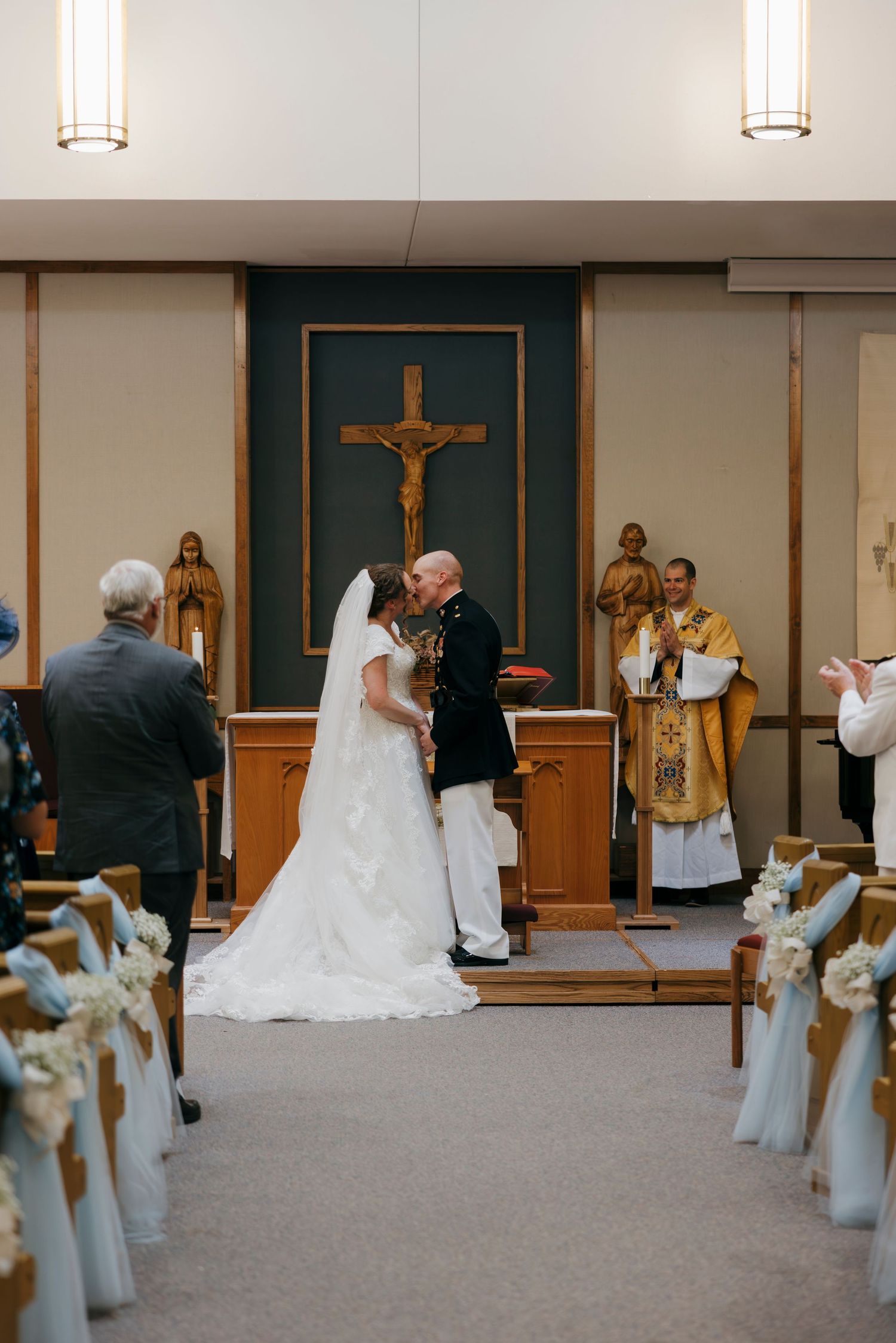 Bride and groom share first kiss at Catholic church altar during wedding ceremony with priest officiating.