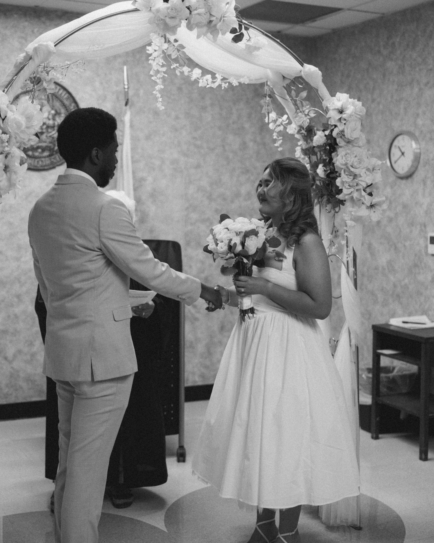 A black and white photo captures an intimate wedding ceremony moment under a decorated arch.