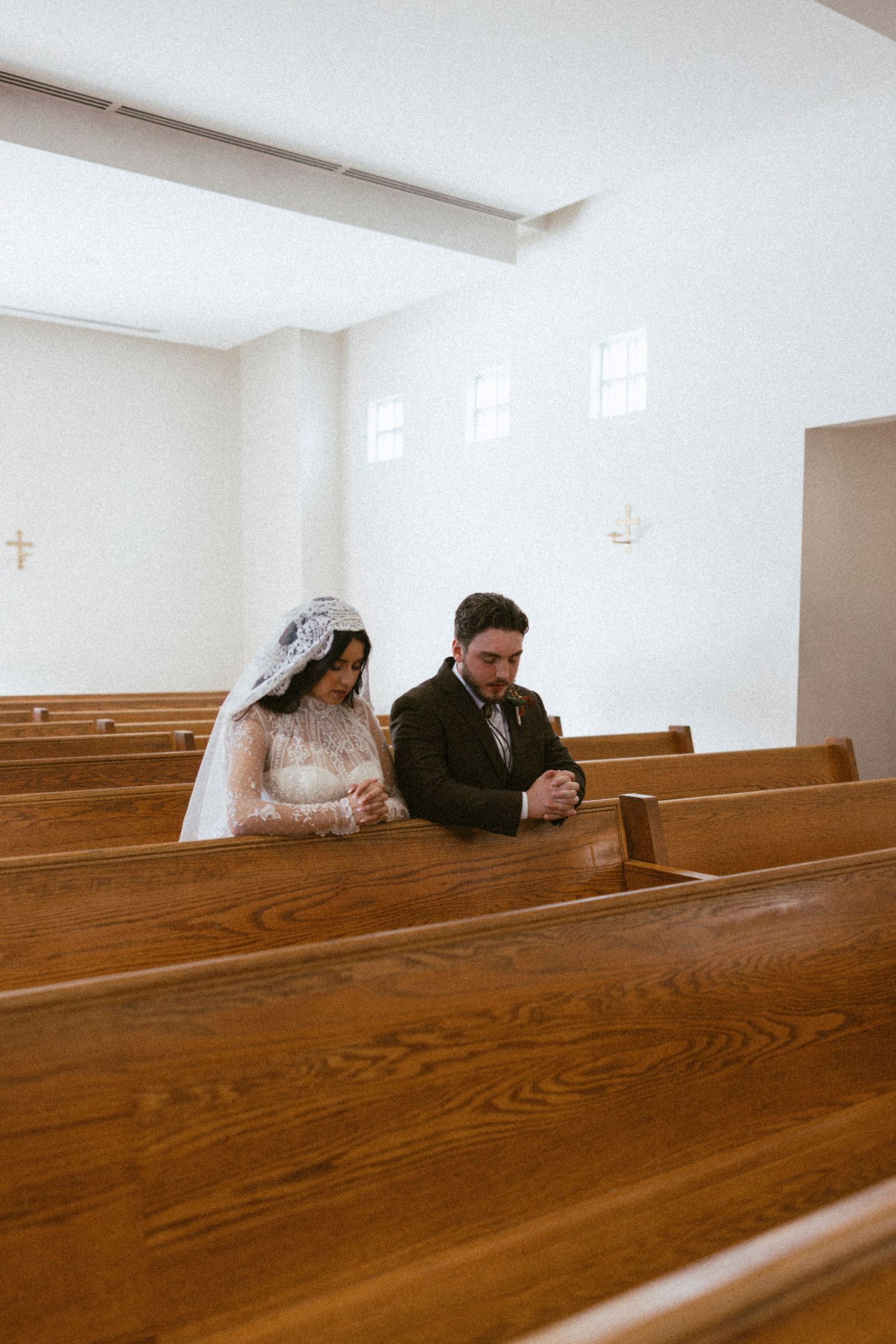 A couple in wedding attire prays together while sitting in a wooden church pew.