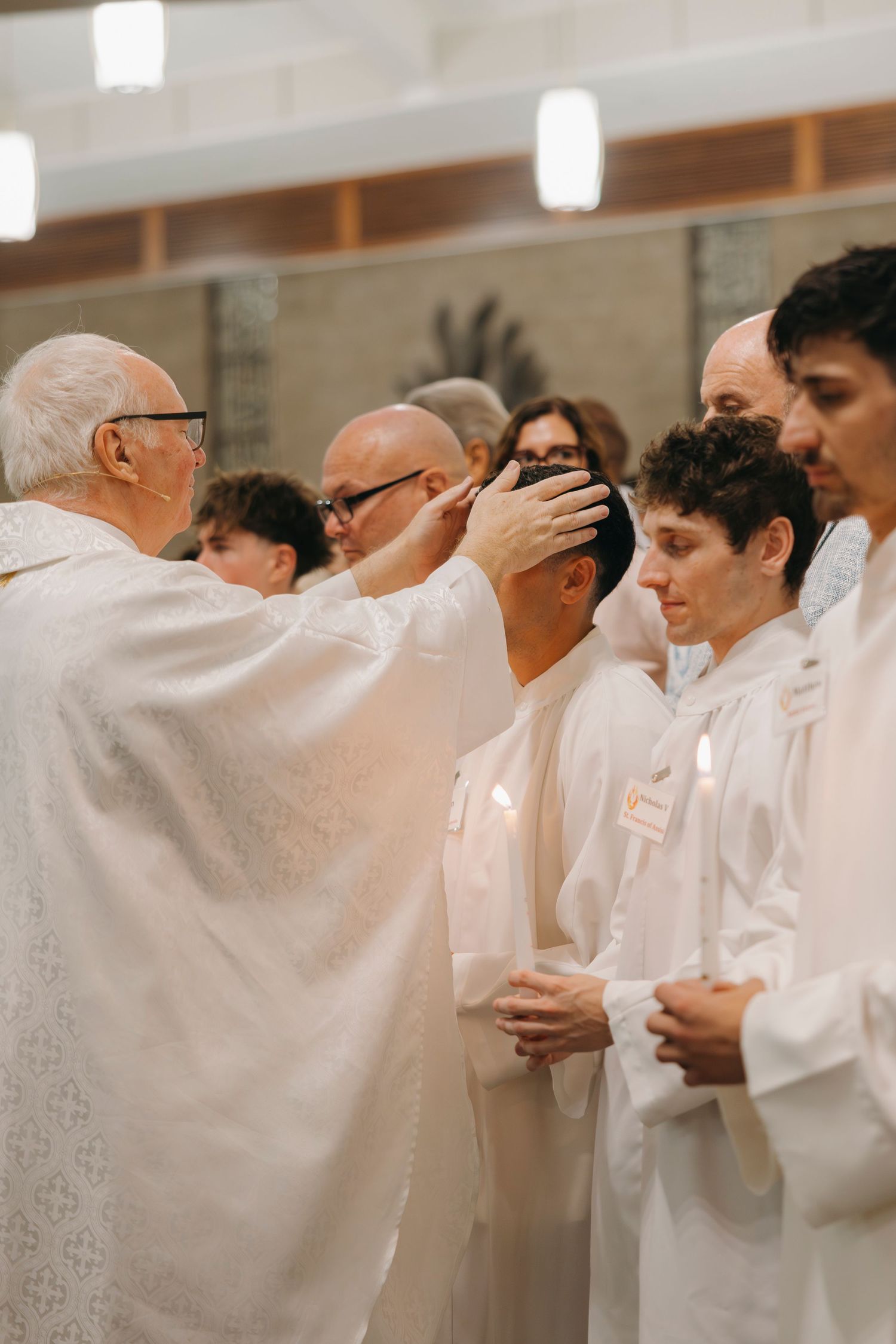 Elderly priest lays hands on candidates in white robes during Confirmation rite at Mass.