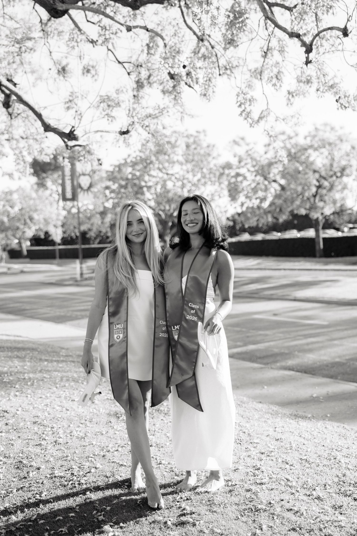 Two women in graduation stoles and white dresses pose outdoors under trees in a black and white photo.