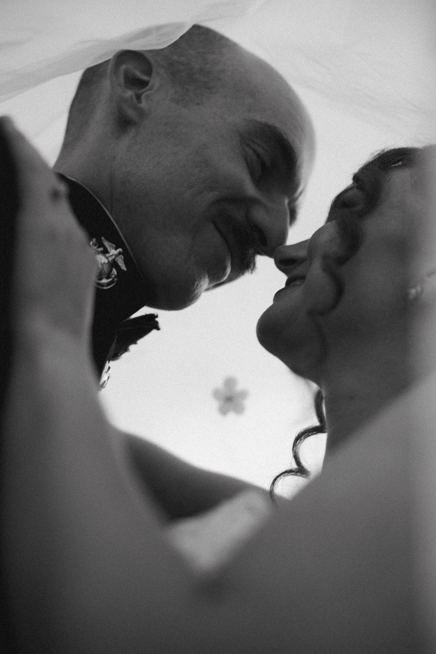 Black and white photo of a couple sharing a romantic kiss under a veil on their wedding day.