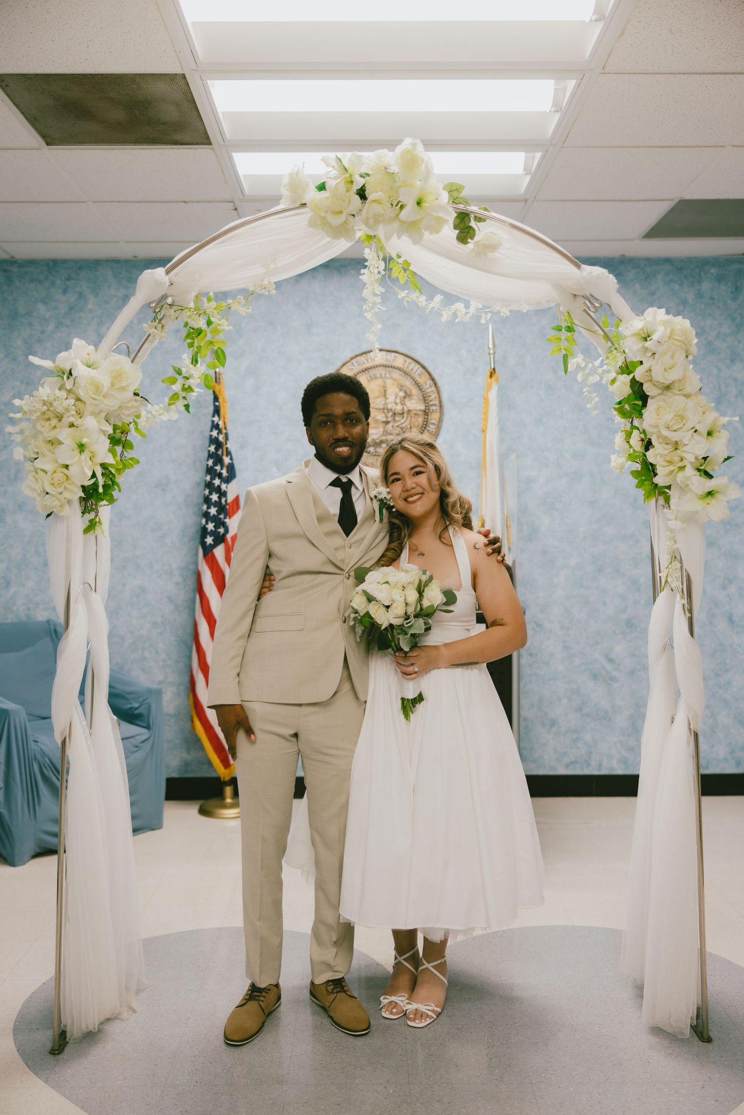 A wedding couple poses under a decorated arch with white flowers and an American flag in the background.