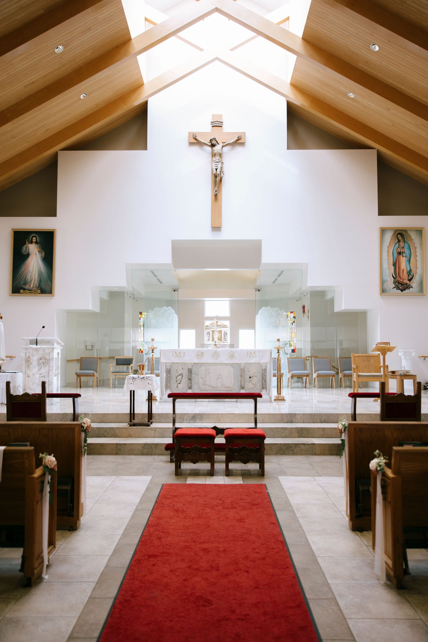Modern church interior with wooden beams, white walls, red carpet aisle, and religious symbols at the altar.