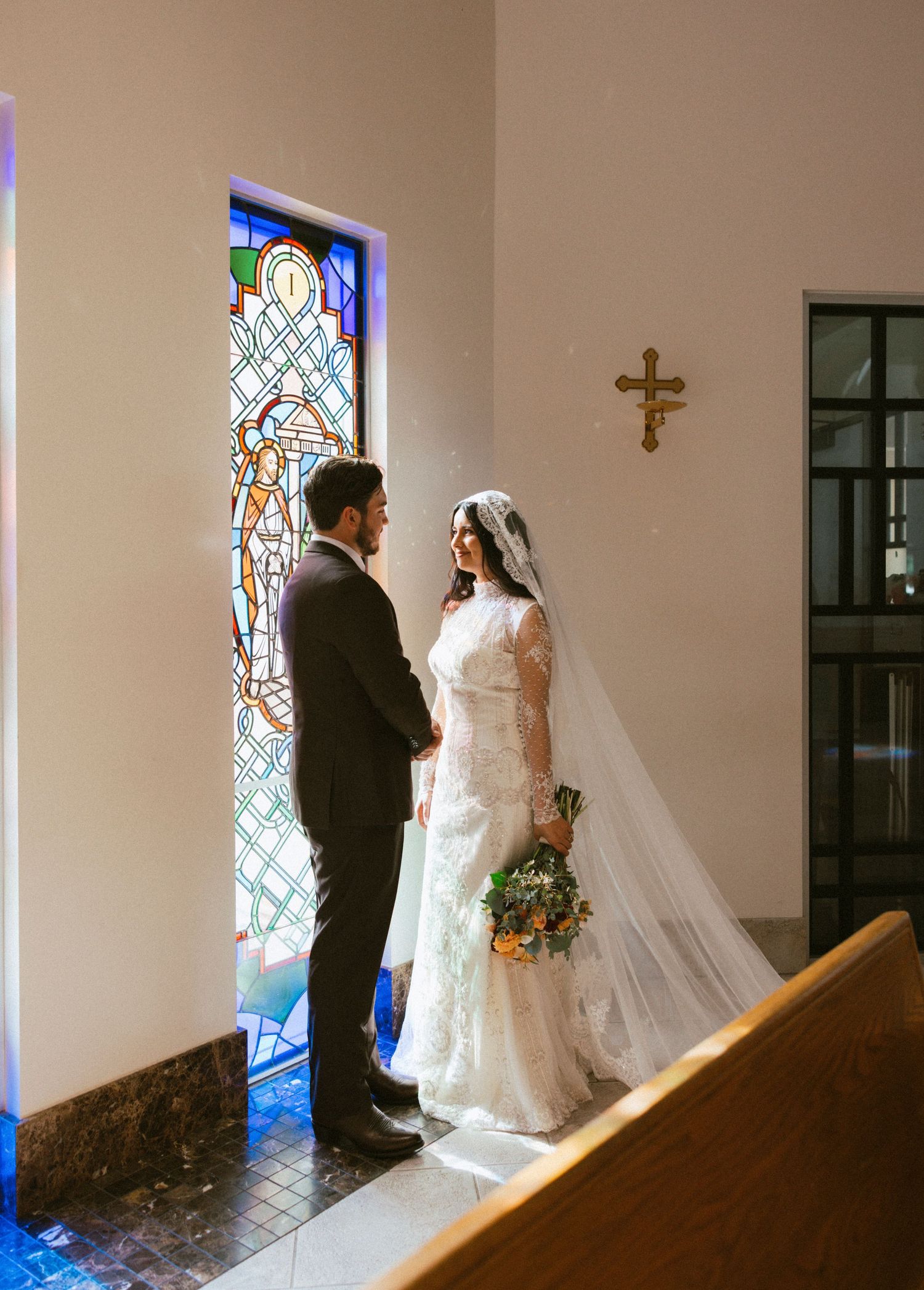 A couple poses in front of colorful stained glass windows in a church setting.