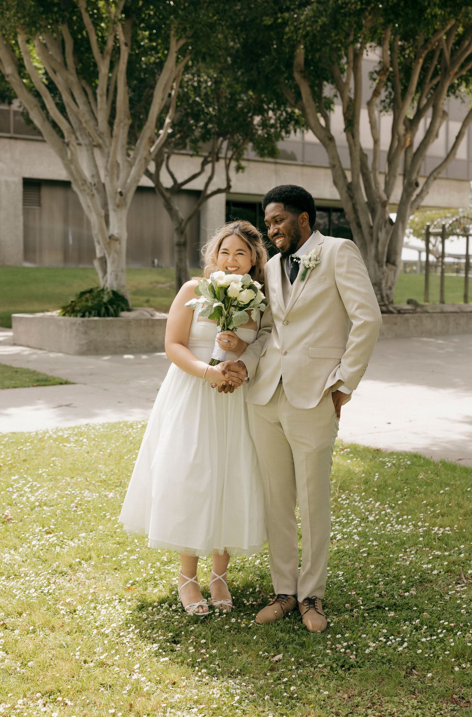 Newlyweds pose together on grass with trees in background, bride holding white bouquet.