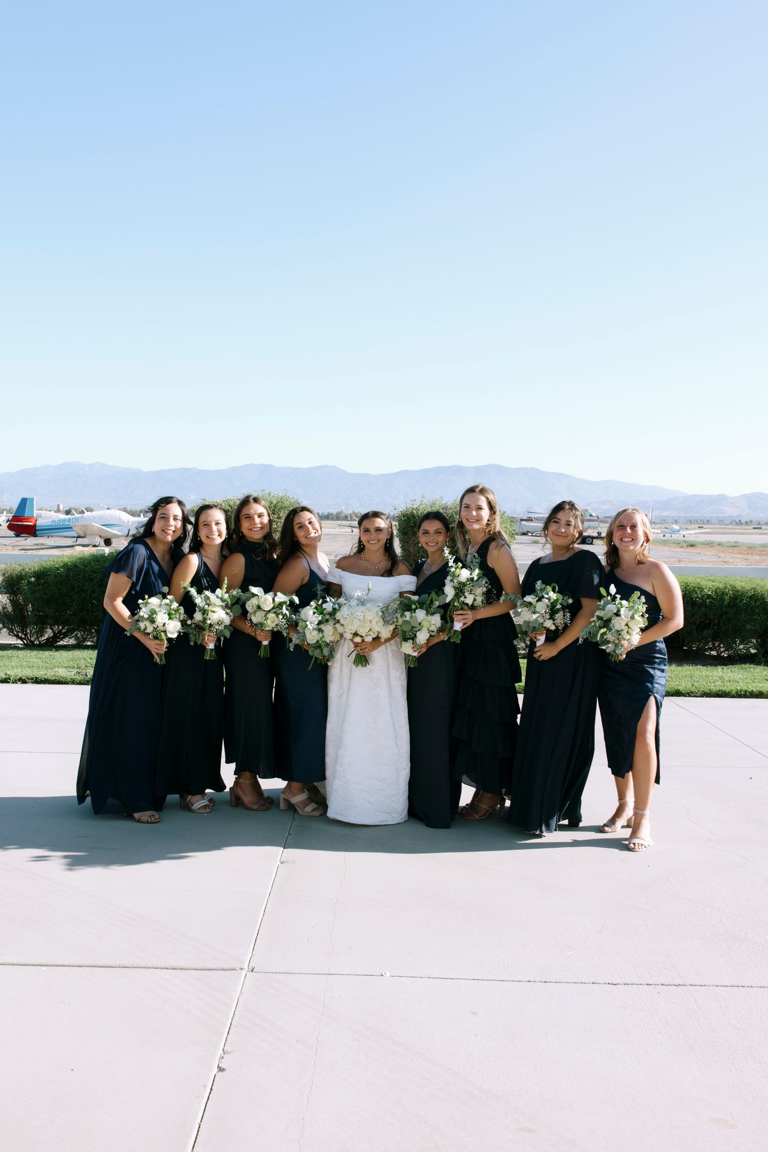 Bridesmaids in black dresses pose with bride in white gown against mountain scenery.