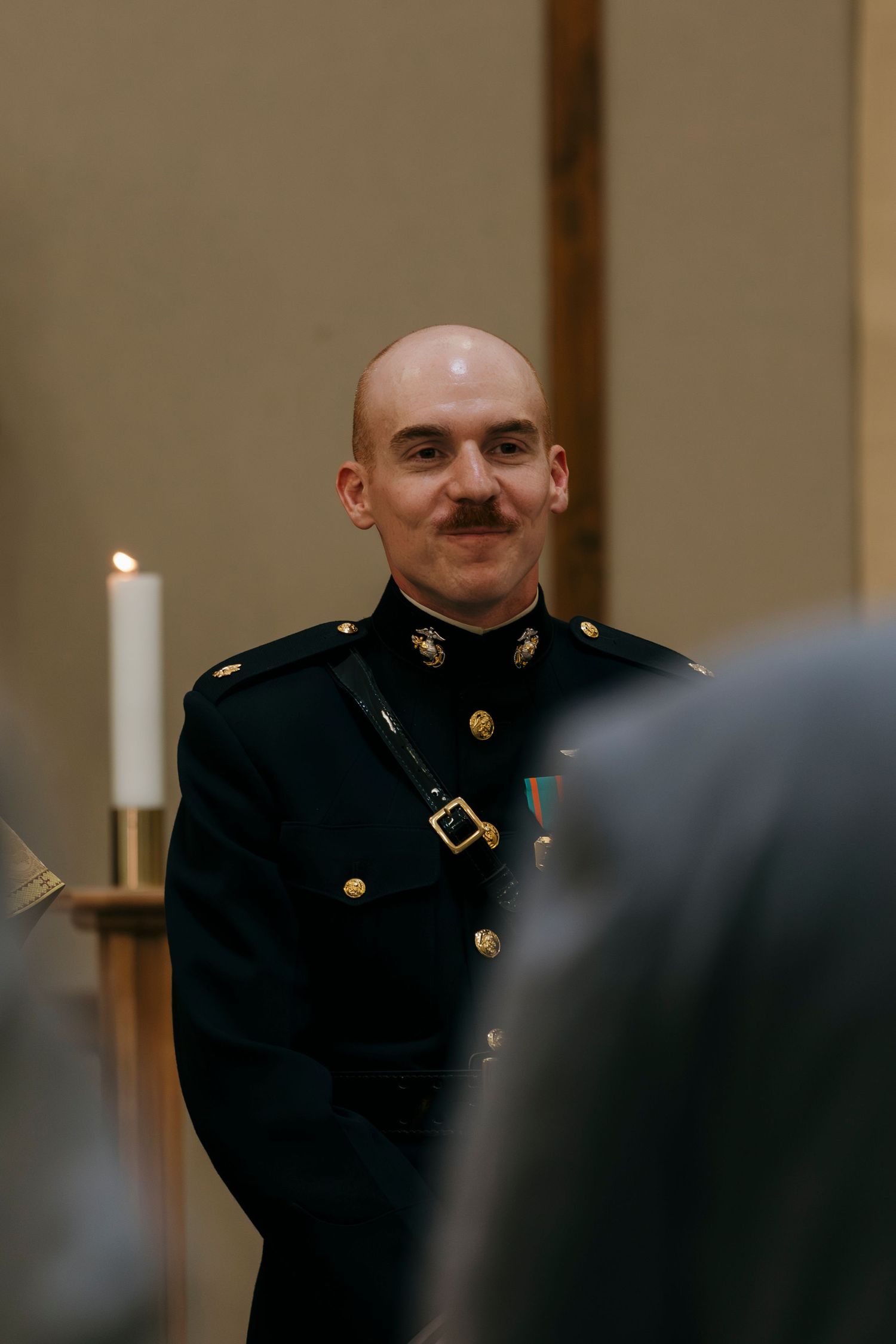 Smiling bald man with mustache in dark military dress uniform with gold buttons and medal at formal ceremony.
