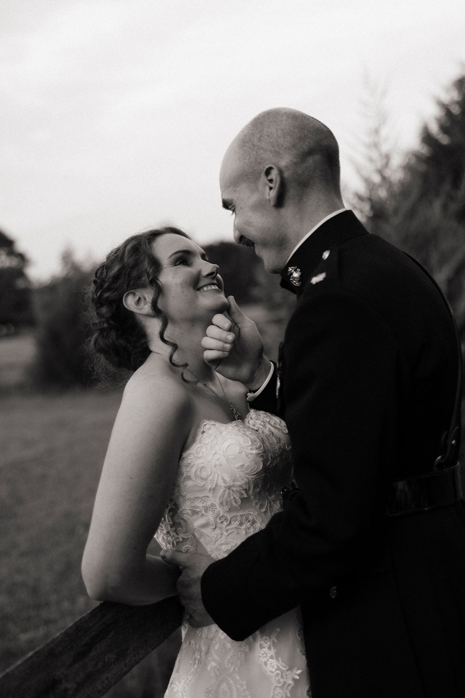 Black and white photo of a smiling bride and groom in military uniform sharing a romantic moment outdoors.