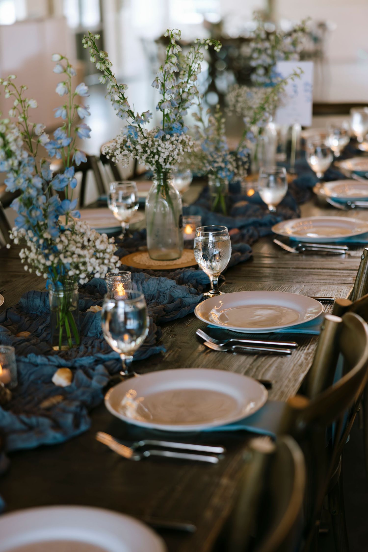 Elegant wedding table setting with blue linen runner, white plates, crystal glasses, and delicate floral centerpieces in glass bottles.