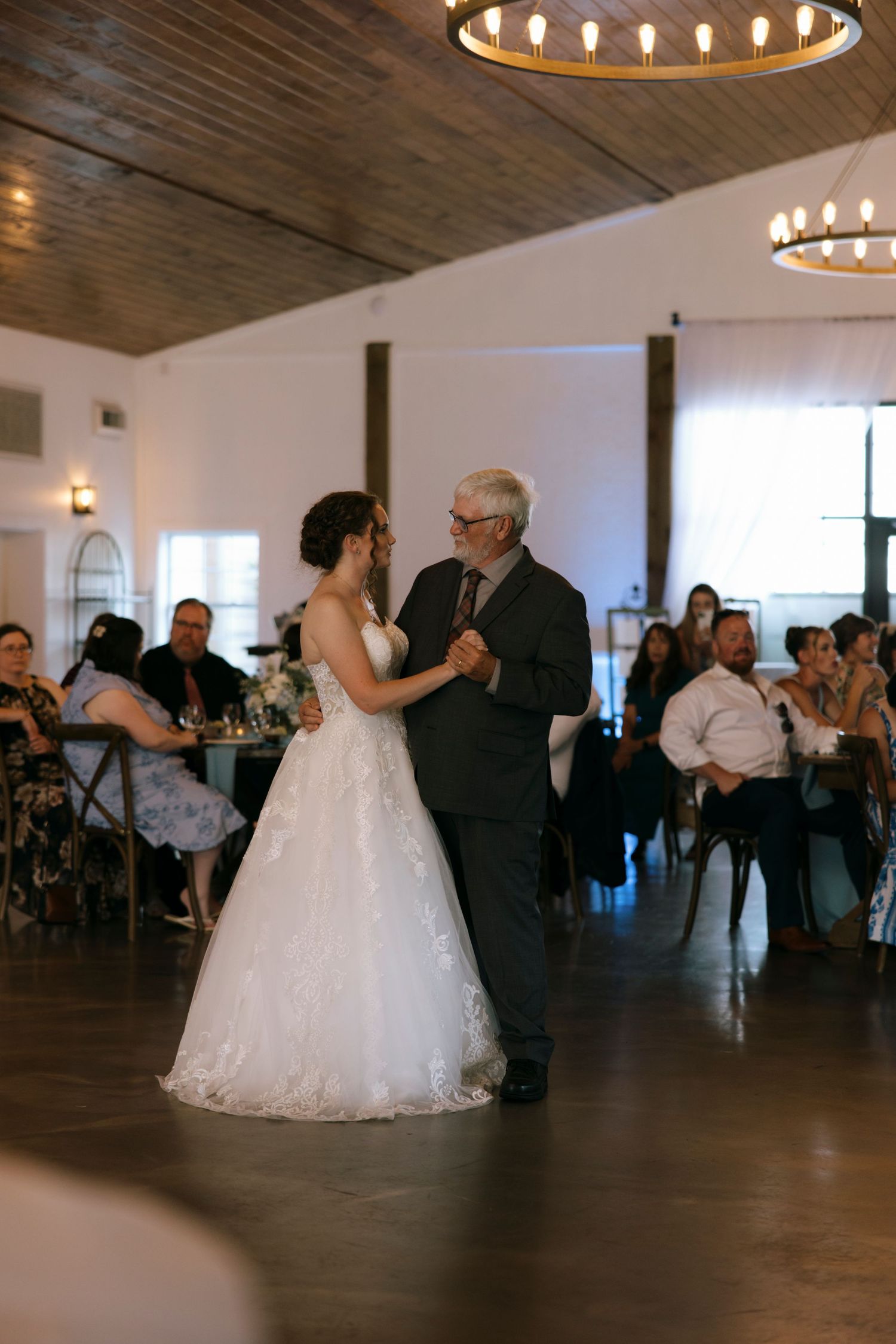 Bride and father share first dance at wedding reception in elegant venue with wood ceiling and chandeliers.
