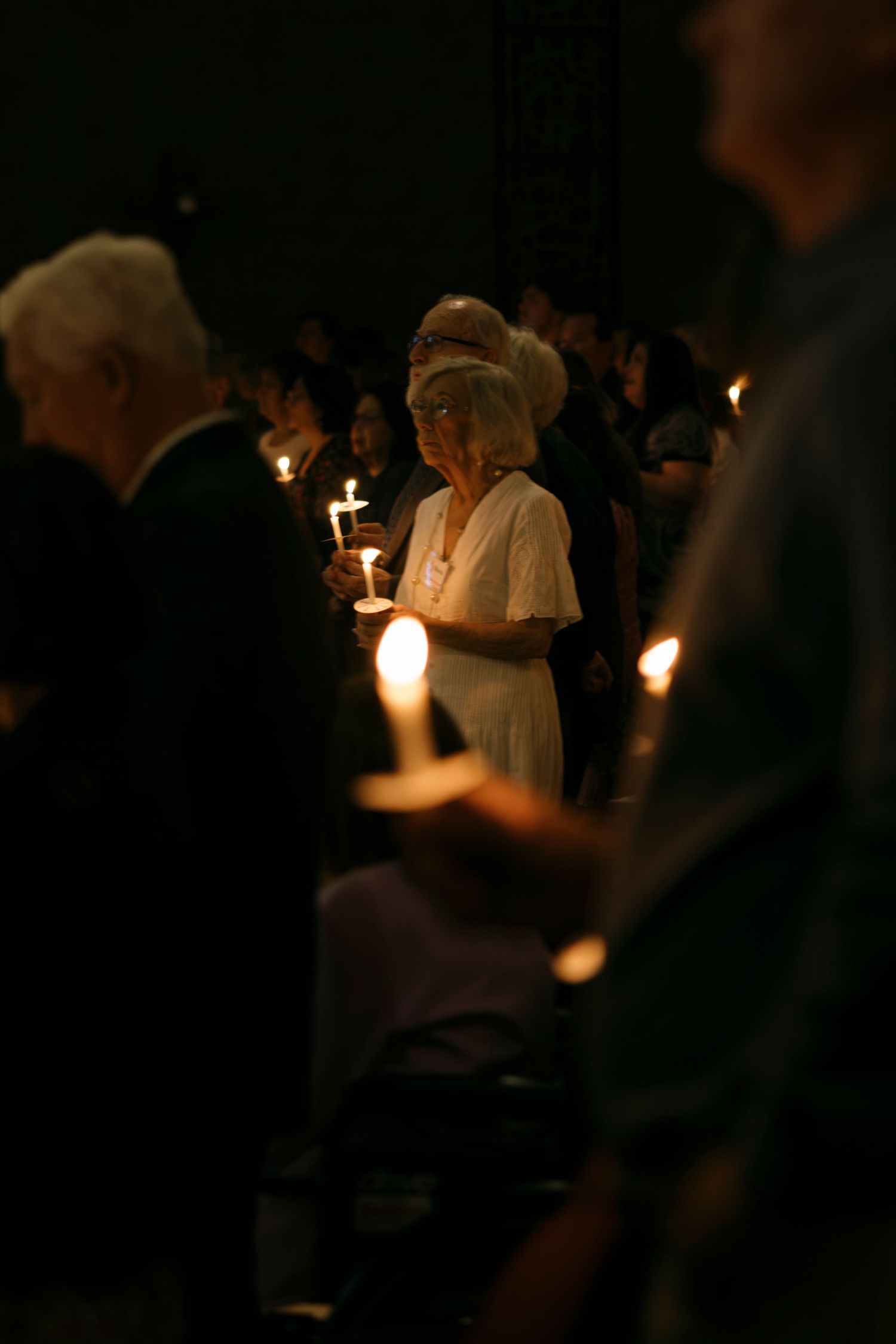 Crowd of people holding candles at a solemn candlelight vigil in a darkened venue.
