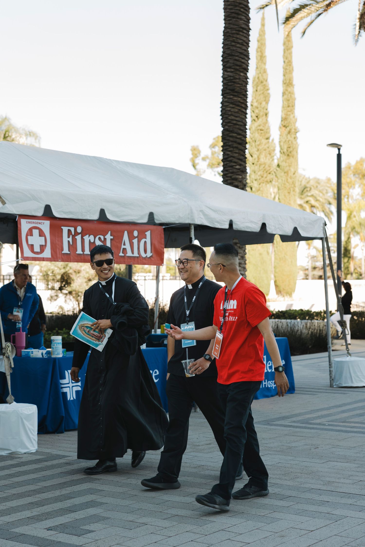 Event staff walk past a First Aid tent at an outdoor venue with palm trees.