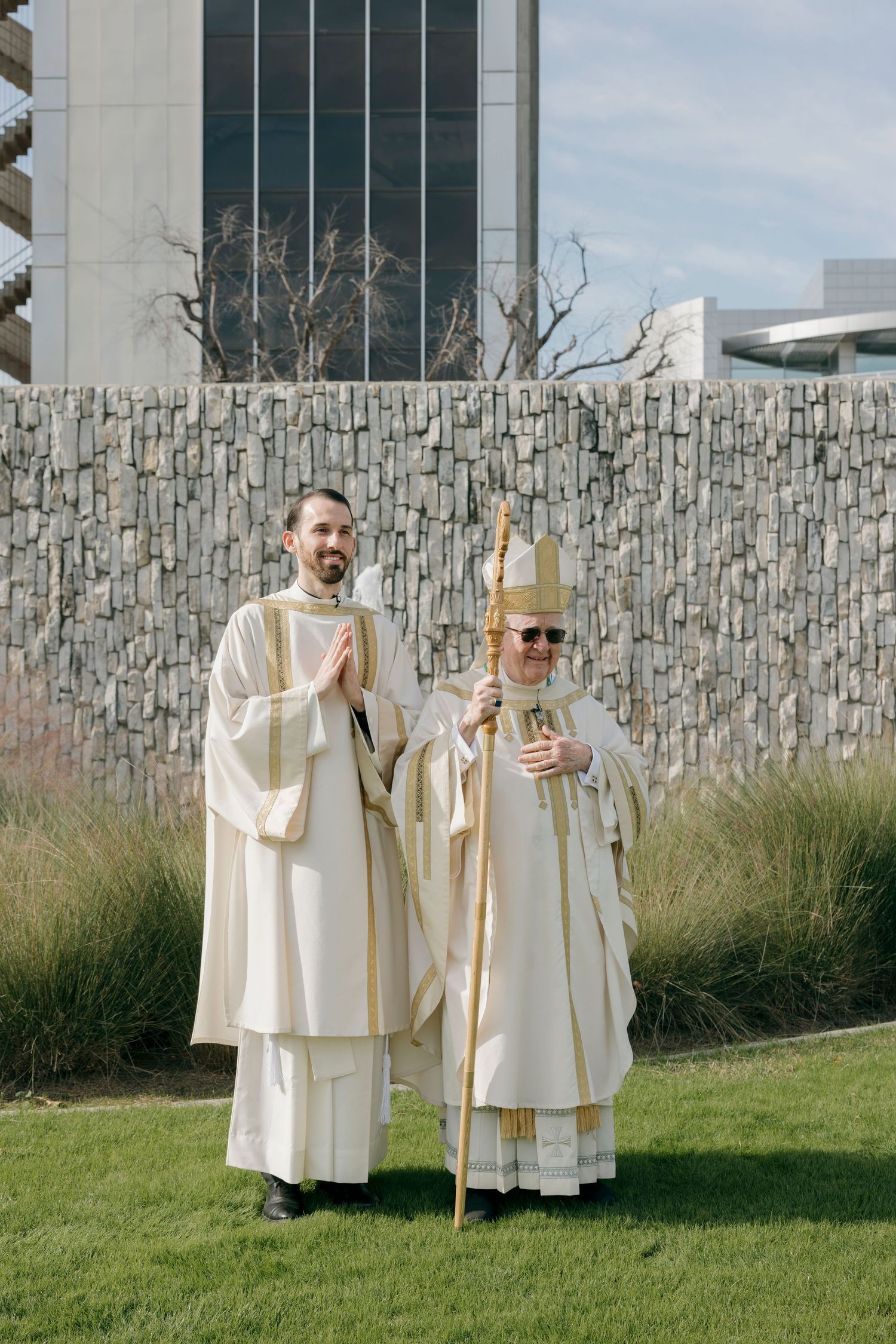 Clergy members in white vestments stand together on a lawn near an architectural wall.