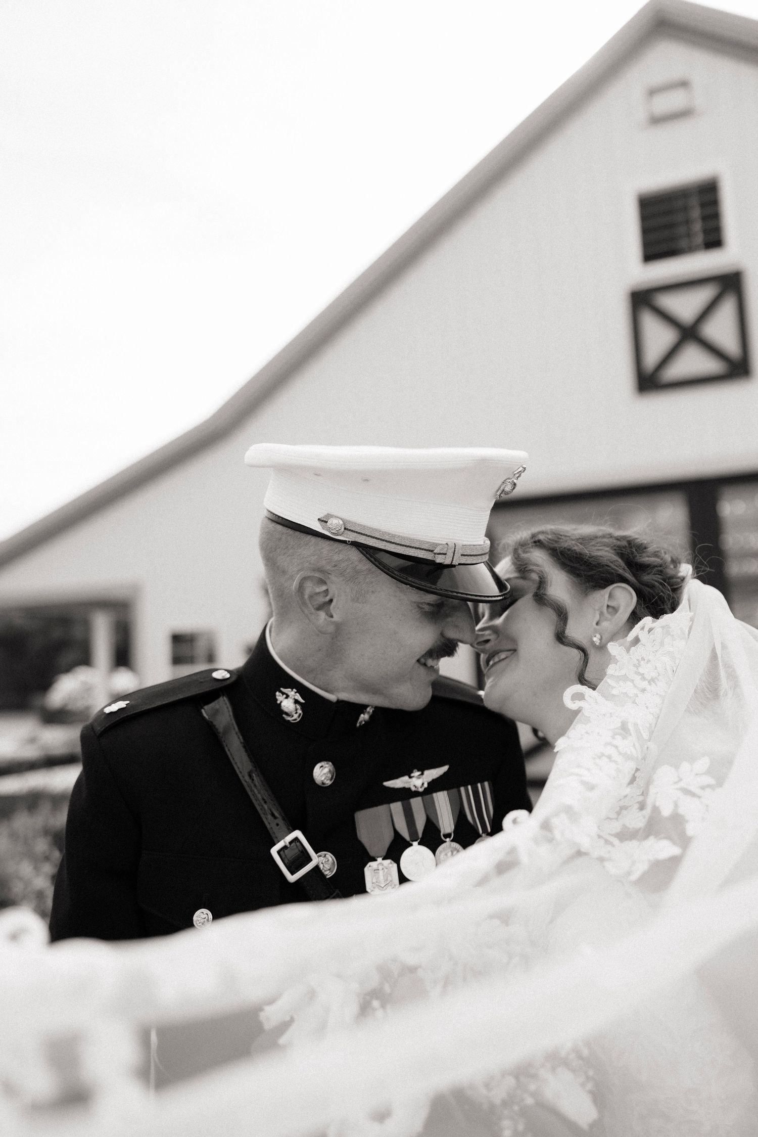 Black and white photo of a Marine in dress uniform sharing a tender moment with his bride in a wedding veil outdoors.