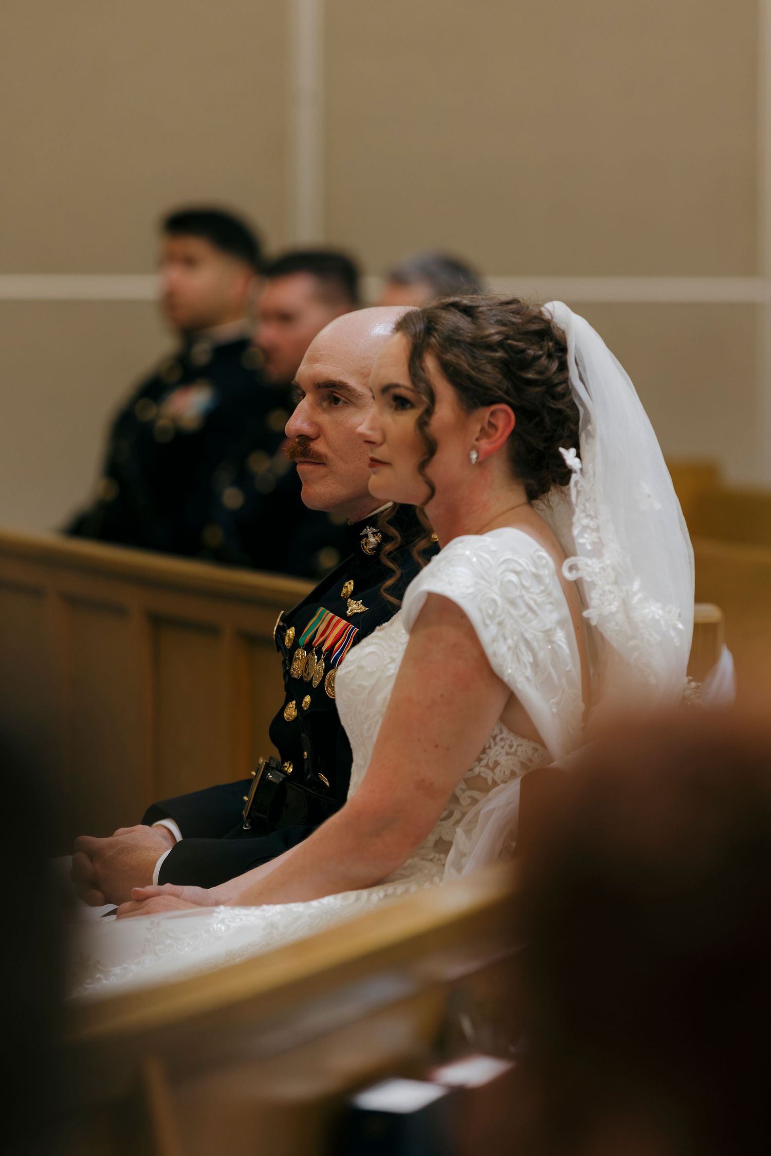 Military groom in dress uniform sits beside bride in white lace gown and veil during wedding ceremony.