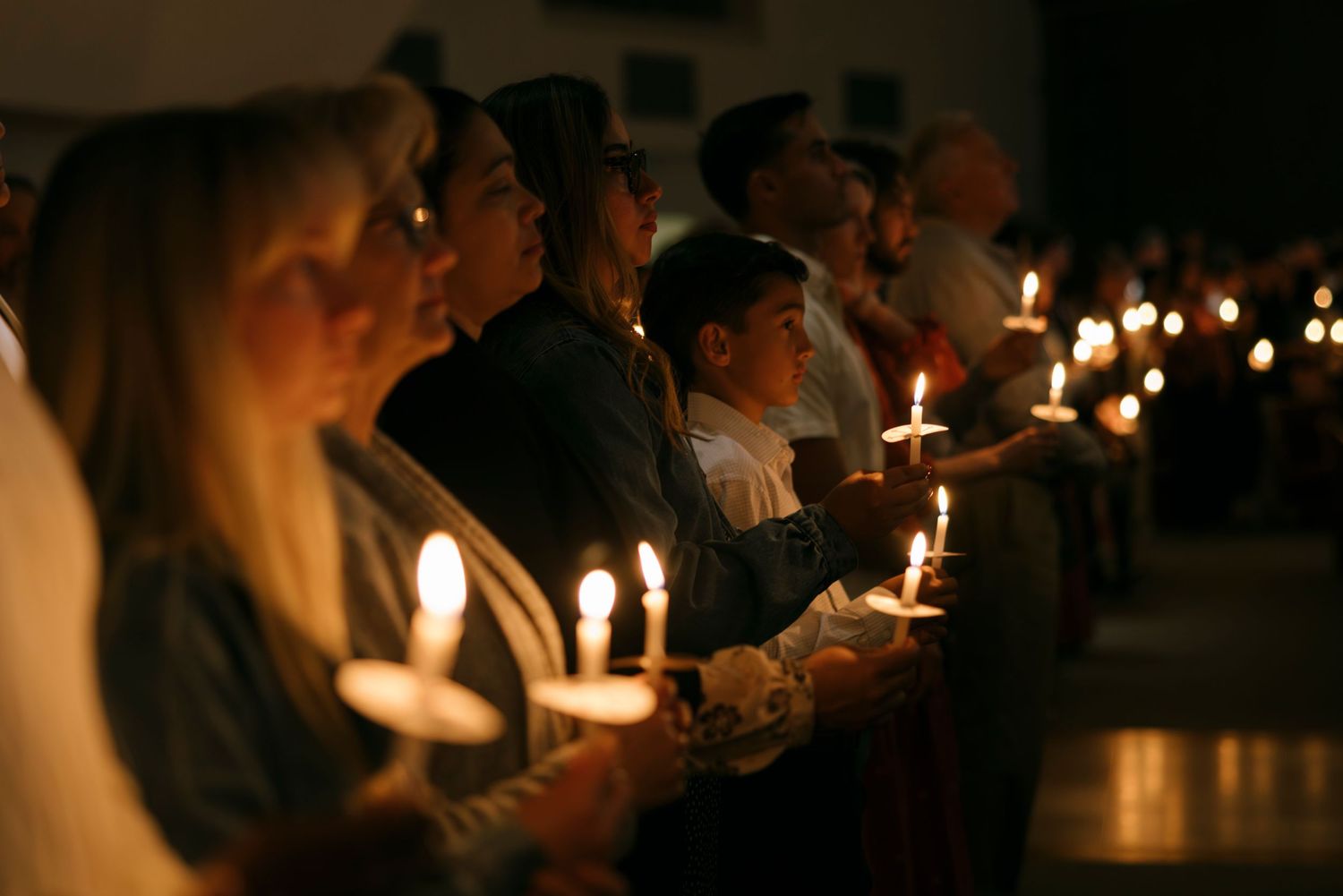 People holding lit candles during a candlelight vigil ceremony in a dimly lit church or gathering hall.
