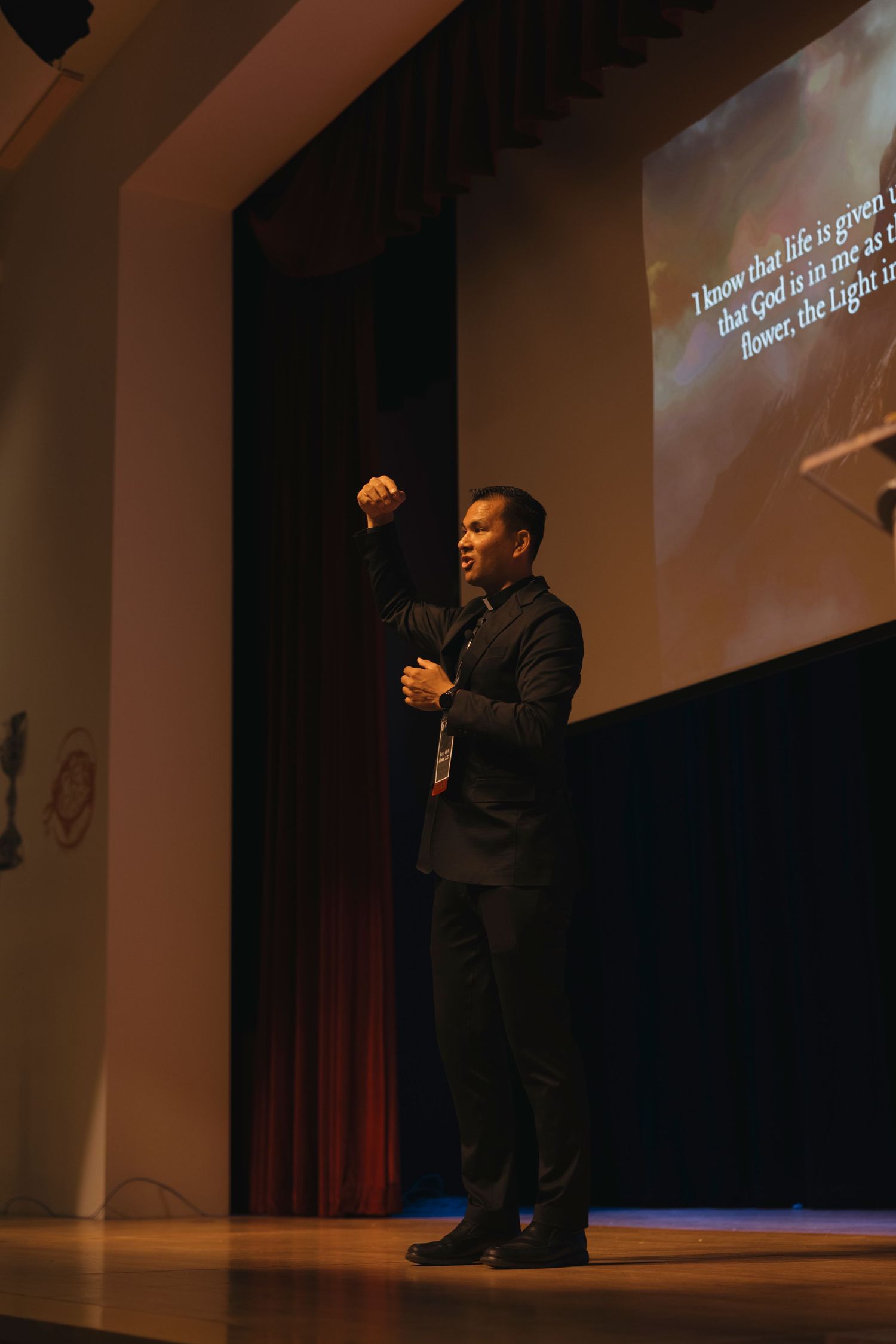 A presenter gestures while speaking on a dimly lit stage with text projected behind them.