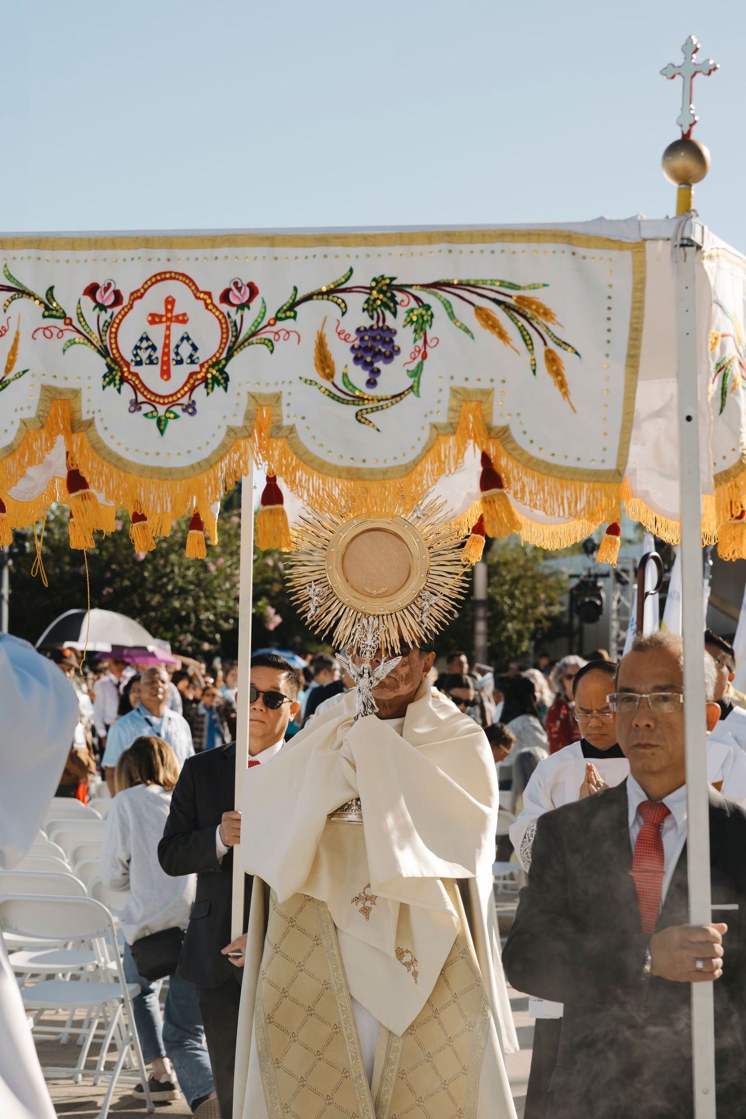 Ornate religious canopy with gold trim and grape motifs carried in outdoor ceremony.