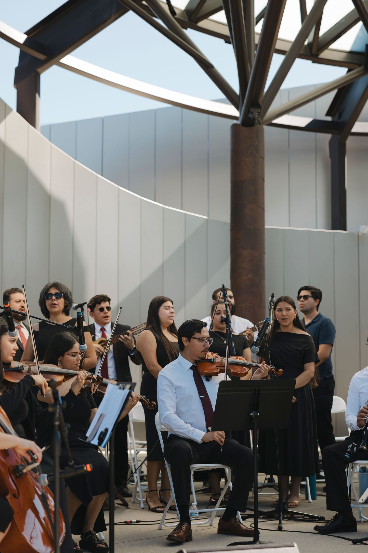 Orchestra performers in formal attire playing instruments under wooden beams.