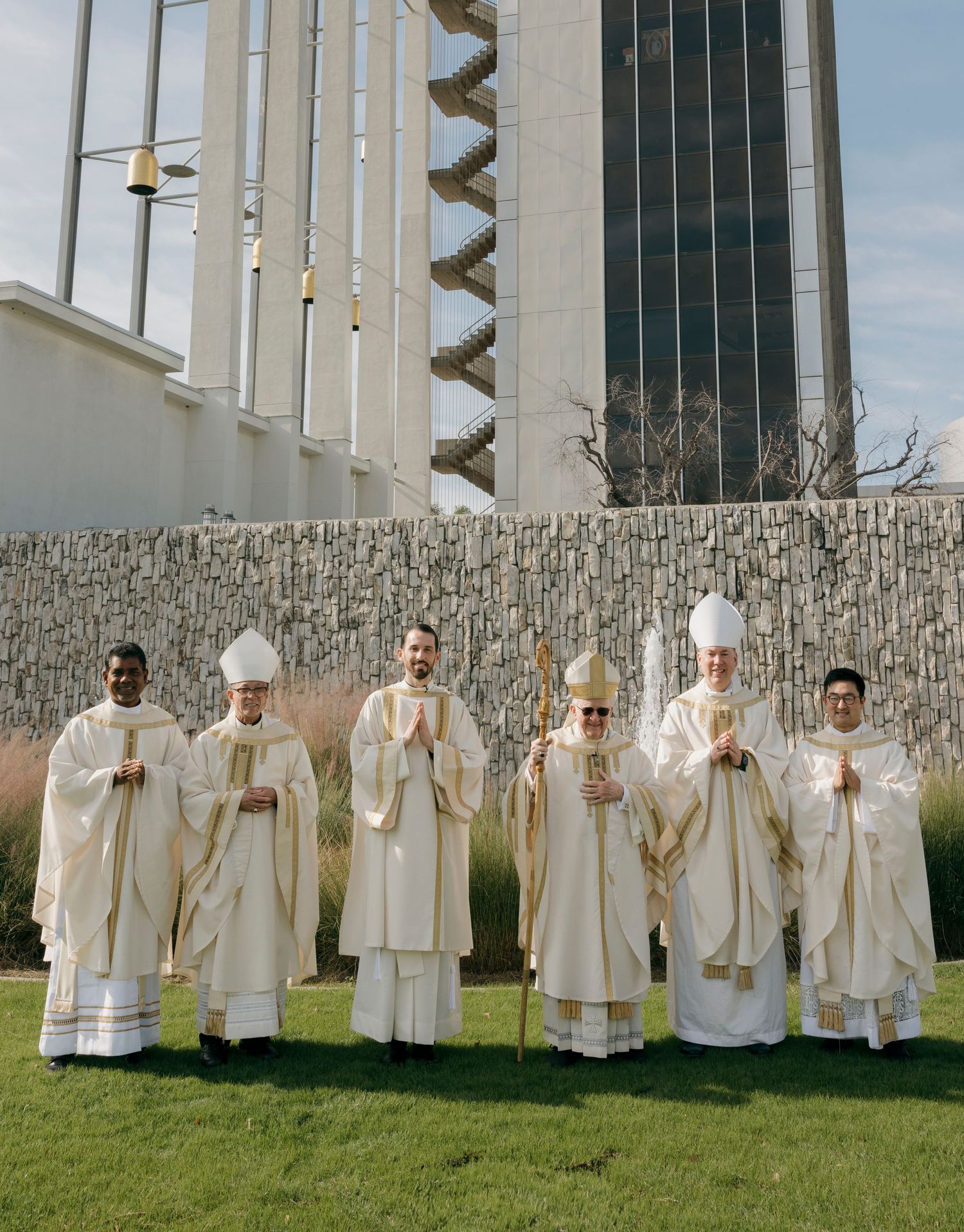Religious leaders in white ceremonial robes gather on grass near a modern building.