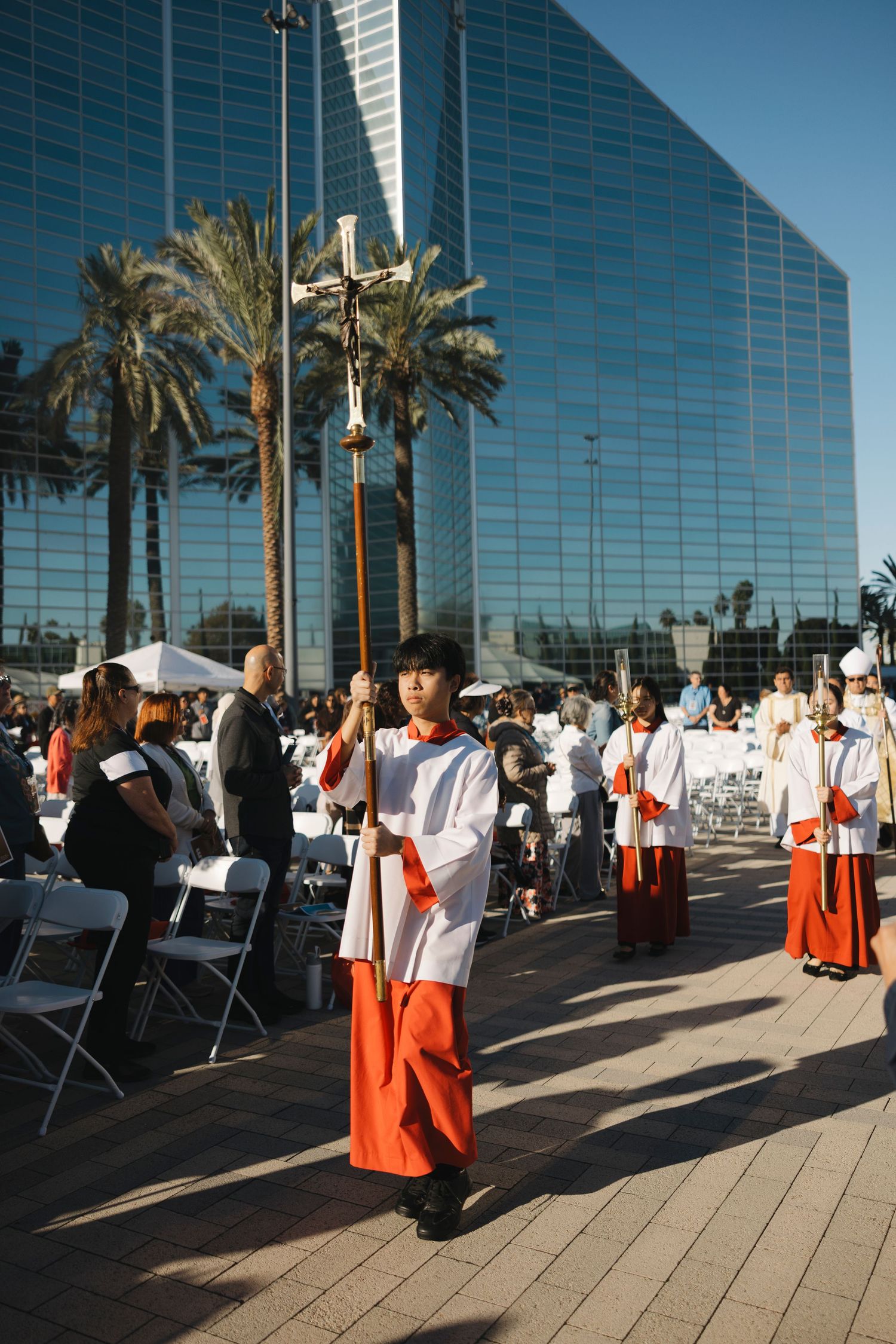 Church procession with clergy and altar servers walking along a modern glass building exterior.