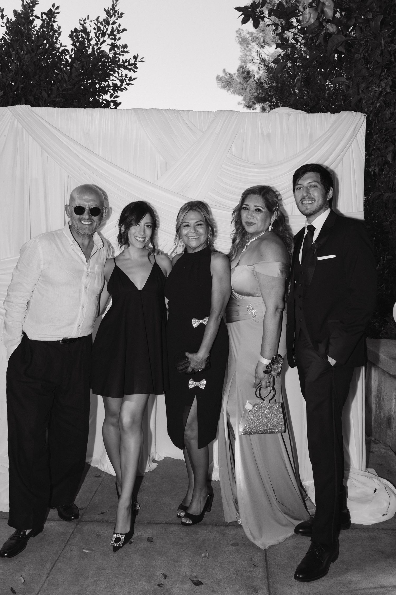 Group of five people posing in formal attire against a white backdrop at an outdoor event.