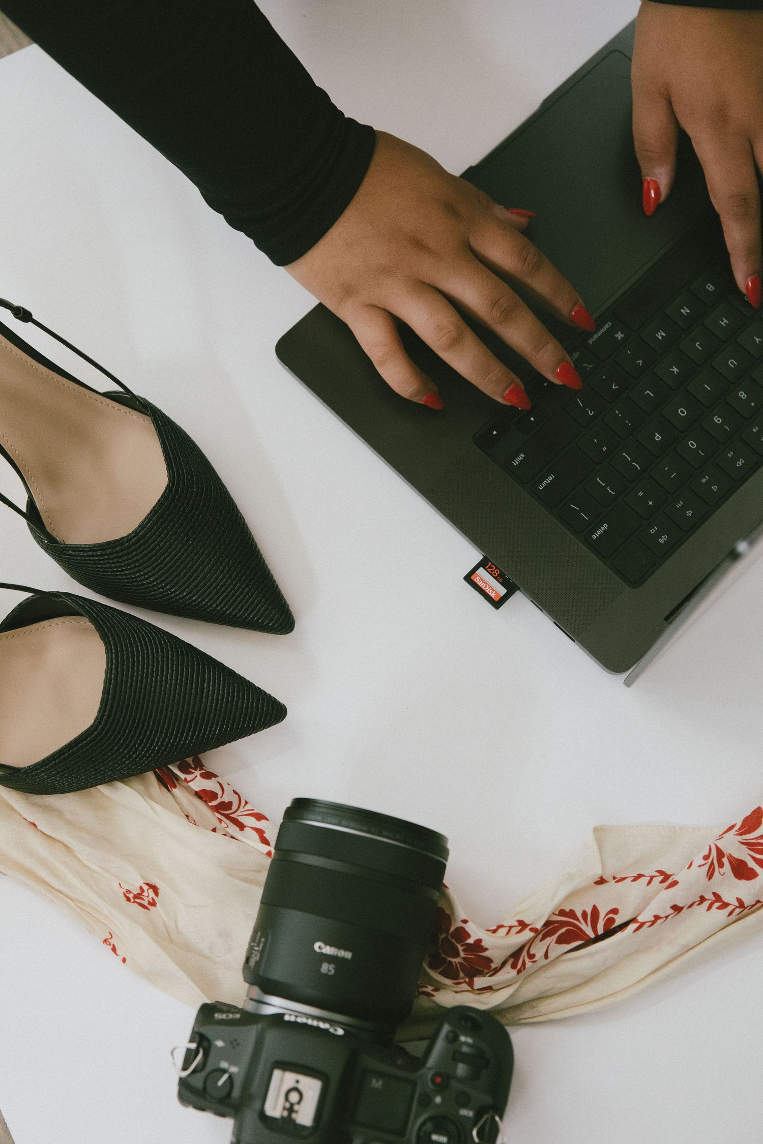 Overhead view of camera, black pointed shoes, and hands typing on laptop keyboard with red nails.