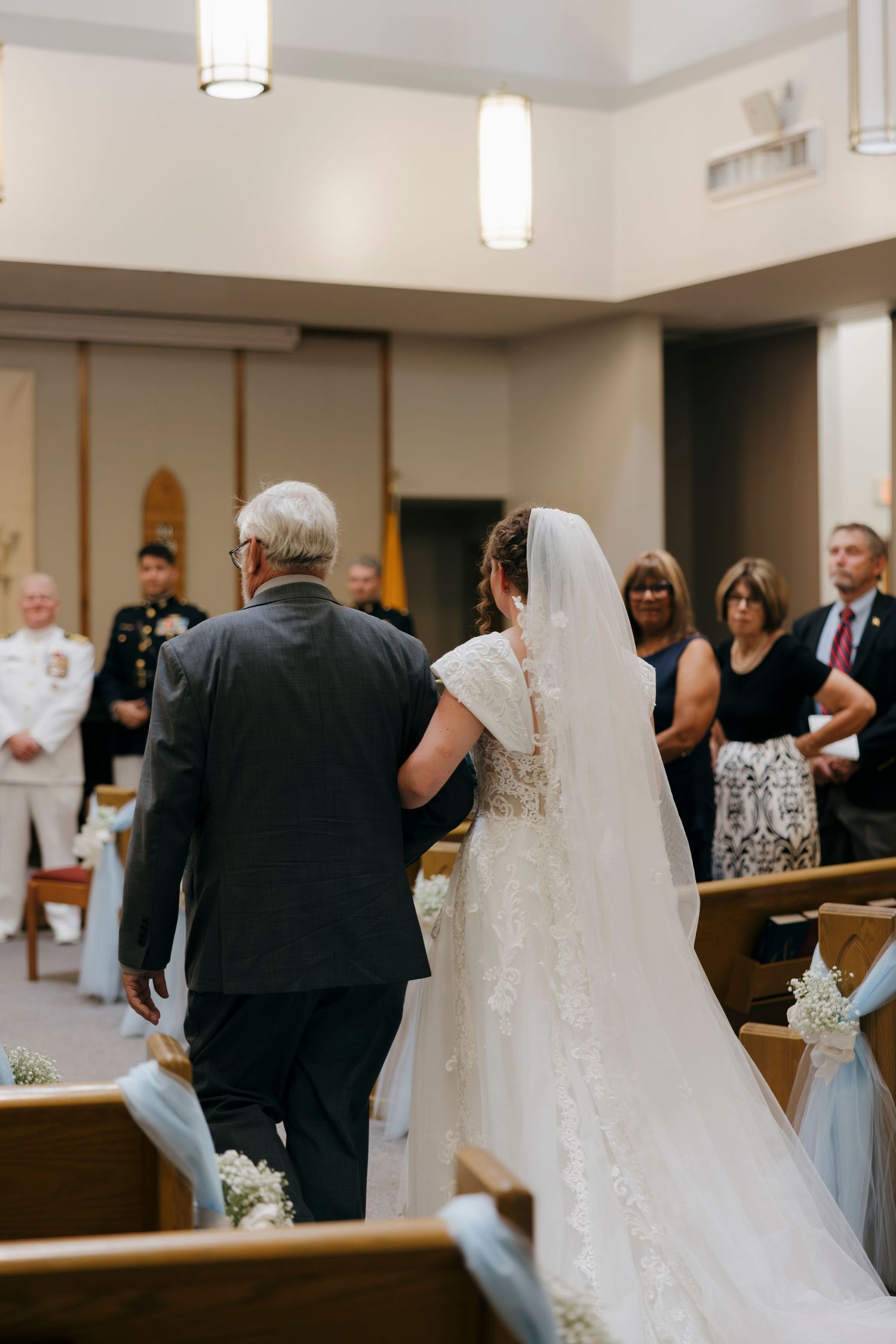 Bride in lace gown and veil walks down church aisle with older man during traditional wedding ceremony.