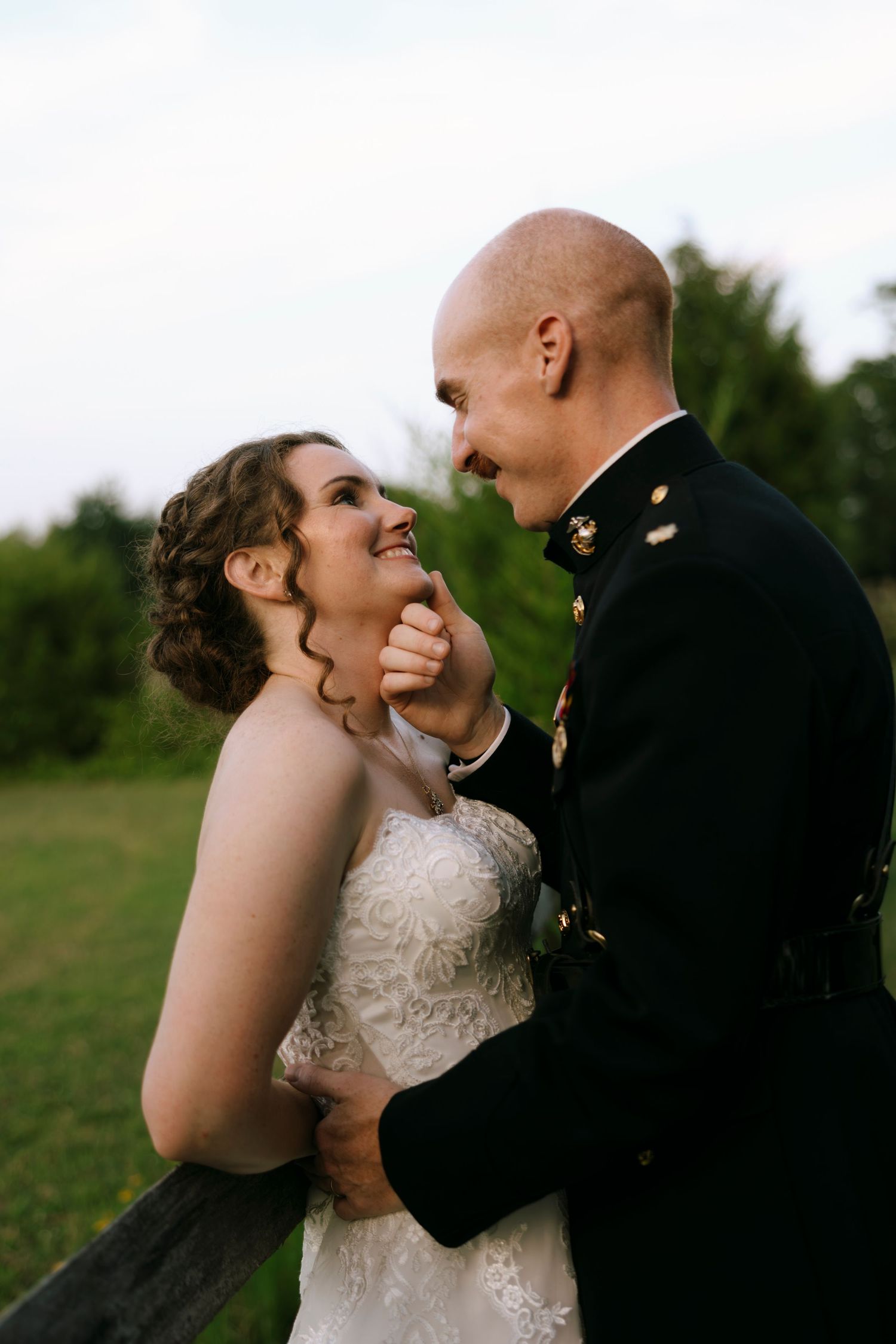 A smiling bride in lace gown and military groom in dress uniform share a romantic moment outdoors.