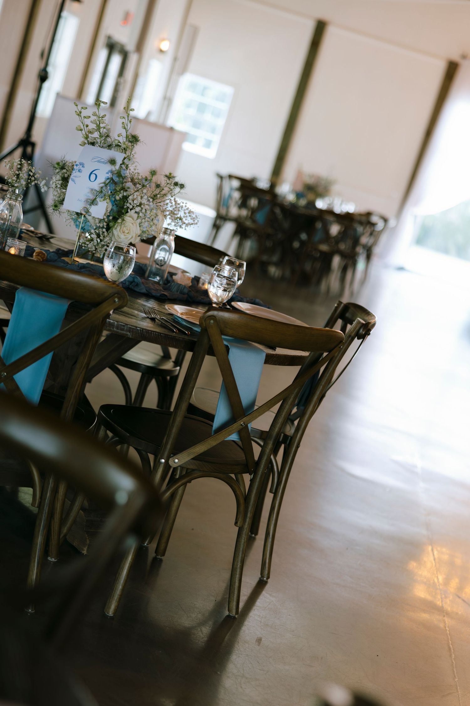 Rustic wedding reception venue with wooden cross-back chairs, teal table runners, floral centerpieces, and elegant glassware on dark tables.