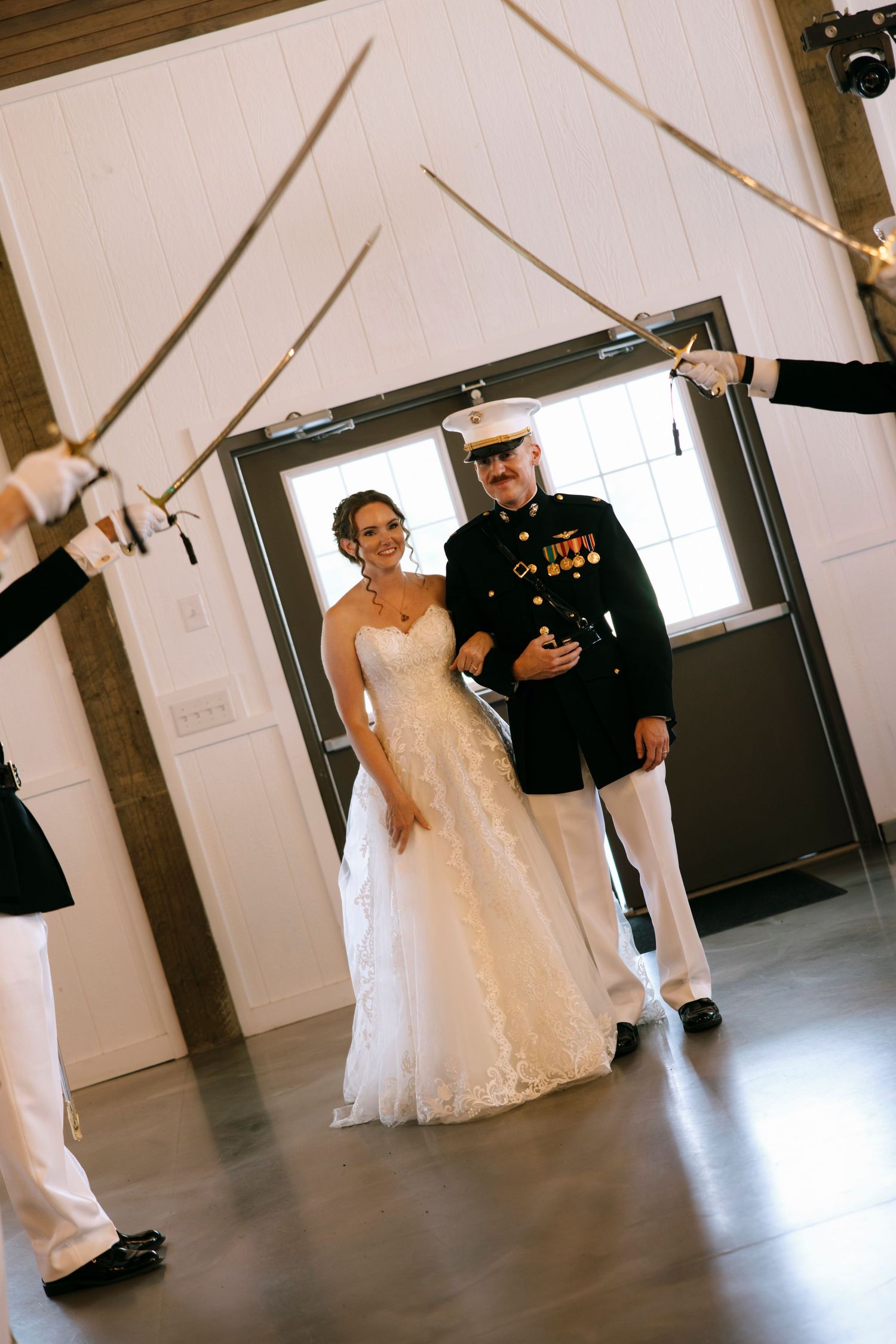 Military wedding couple walks through a sword arch ceremony, bride in white lace gown, groom in dress uniform with medals.