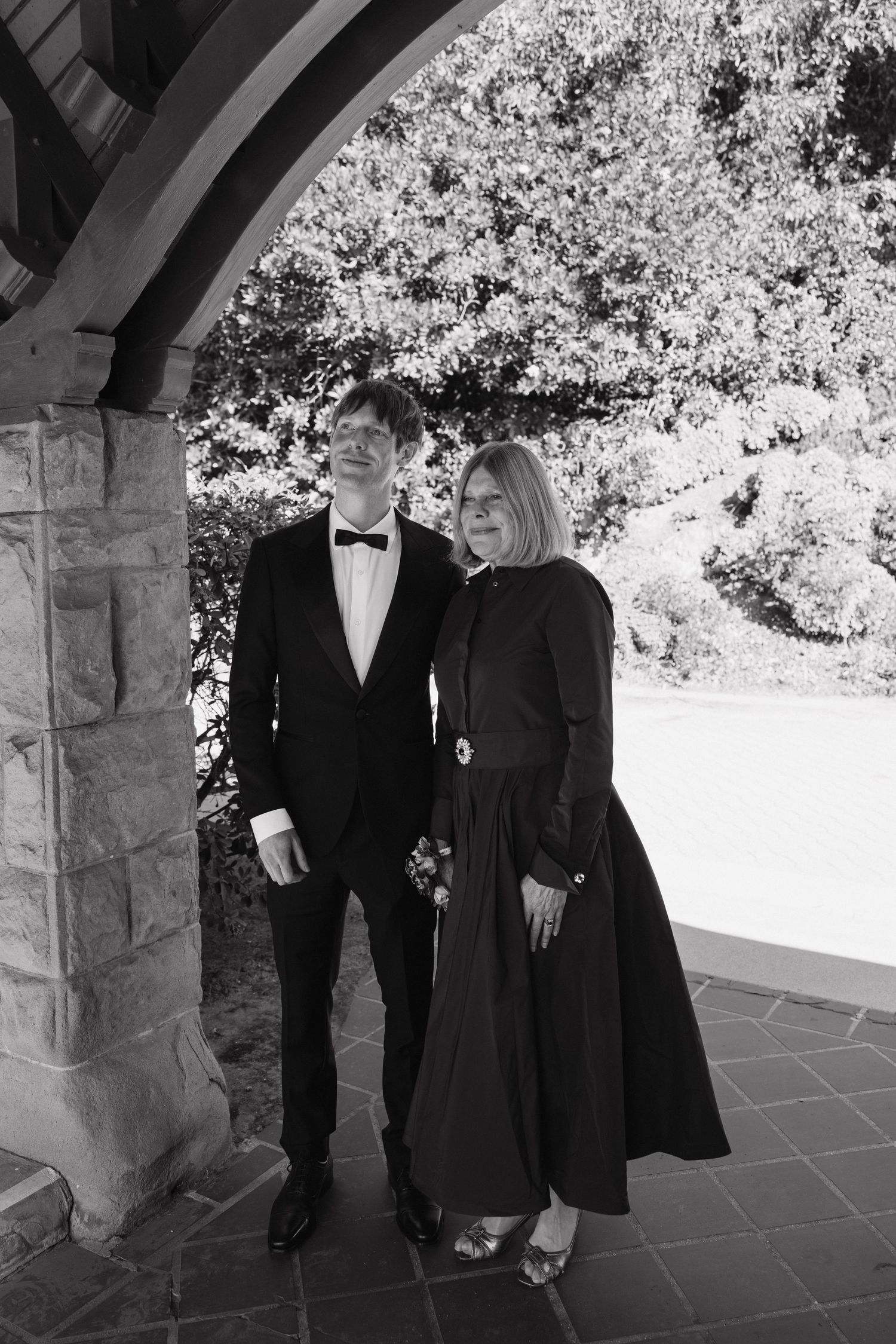 Formal portrait of wedding party members in black attire standing under stone archway.