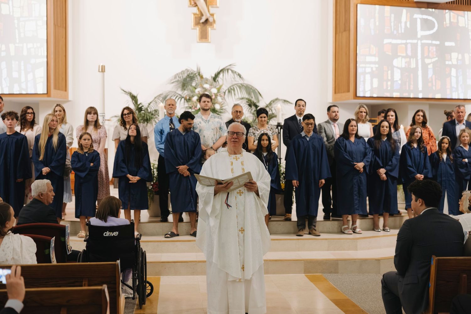 A priest in white robes leads a graduation ceremony in a church with students in blue gowns standing behind him.
