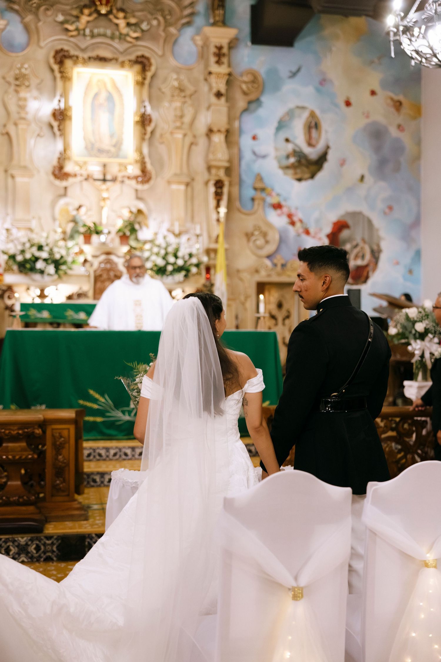 Wedding ceremony in an ornate church with decorative ceiling and white floral arrangements.