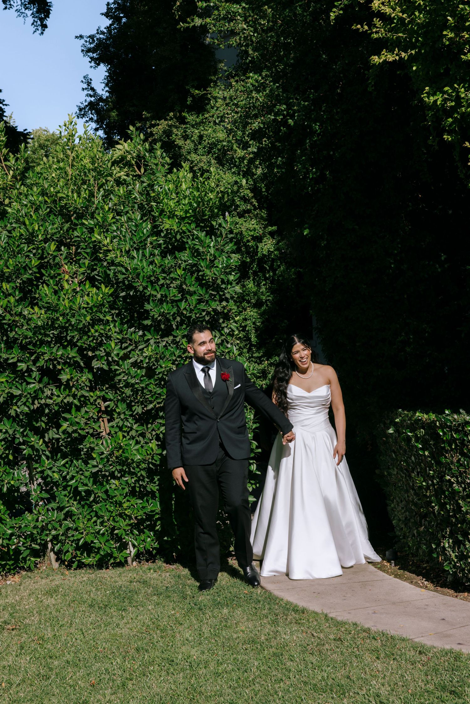 A bride and groom walk hand in hand along a garden path surrounded by lush green hedges on their wedding day.