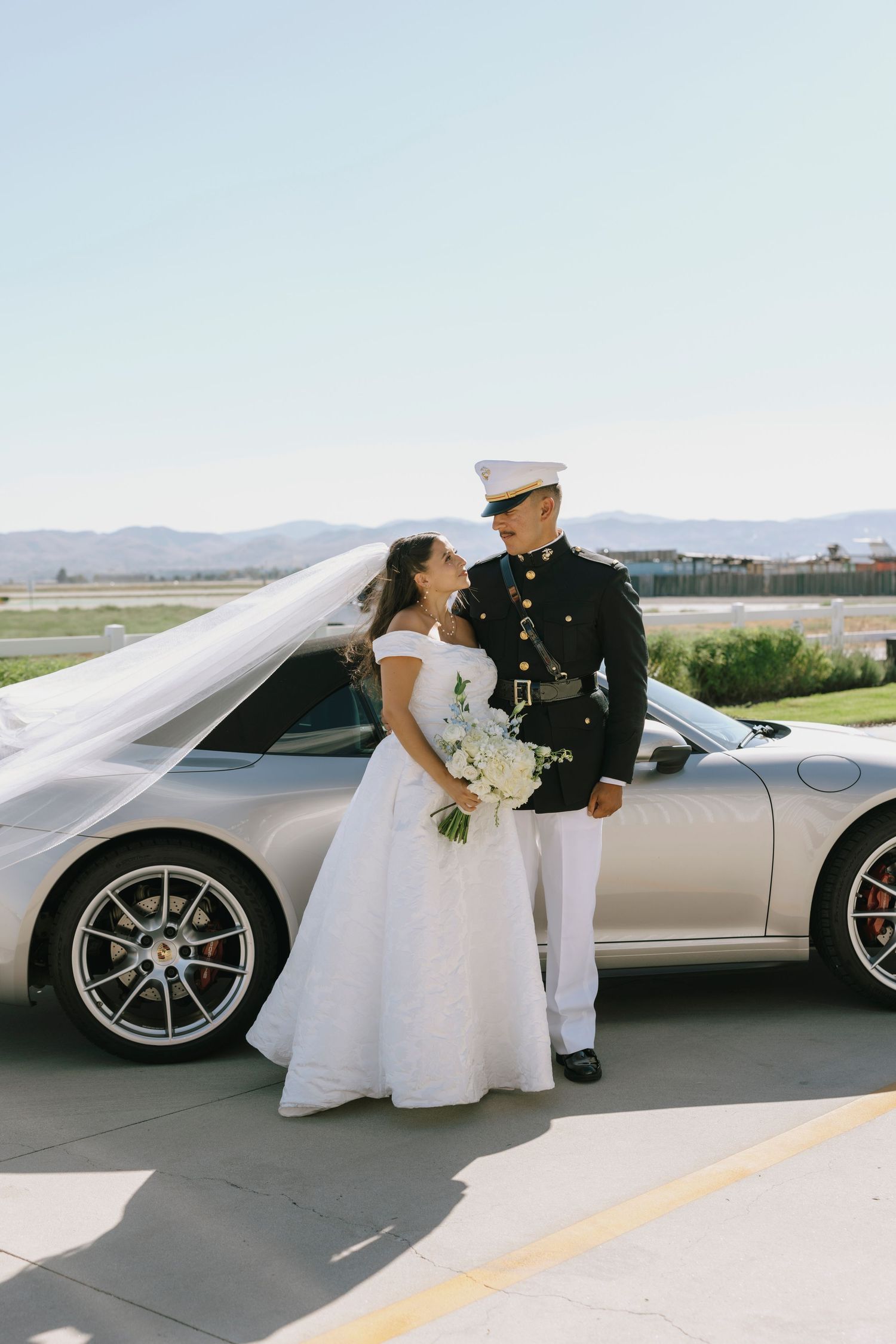 Newlyweds in military dress and wedding gown stand beside a luxury vehicle.