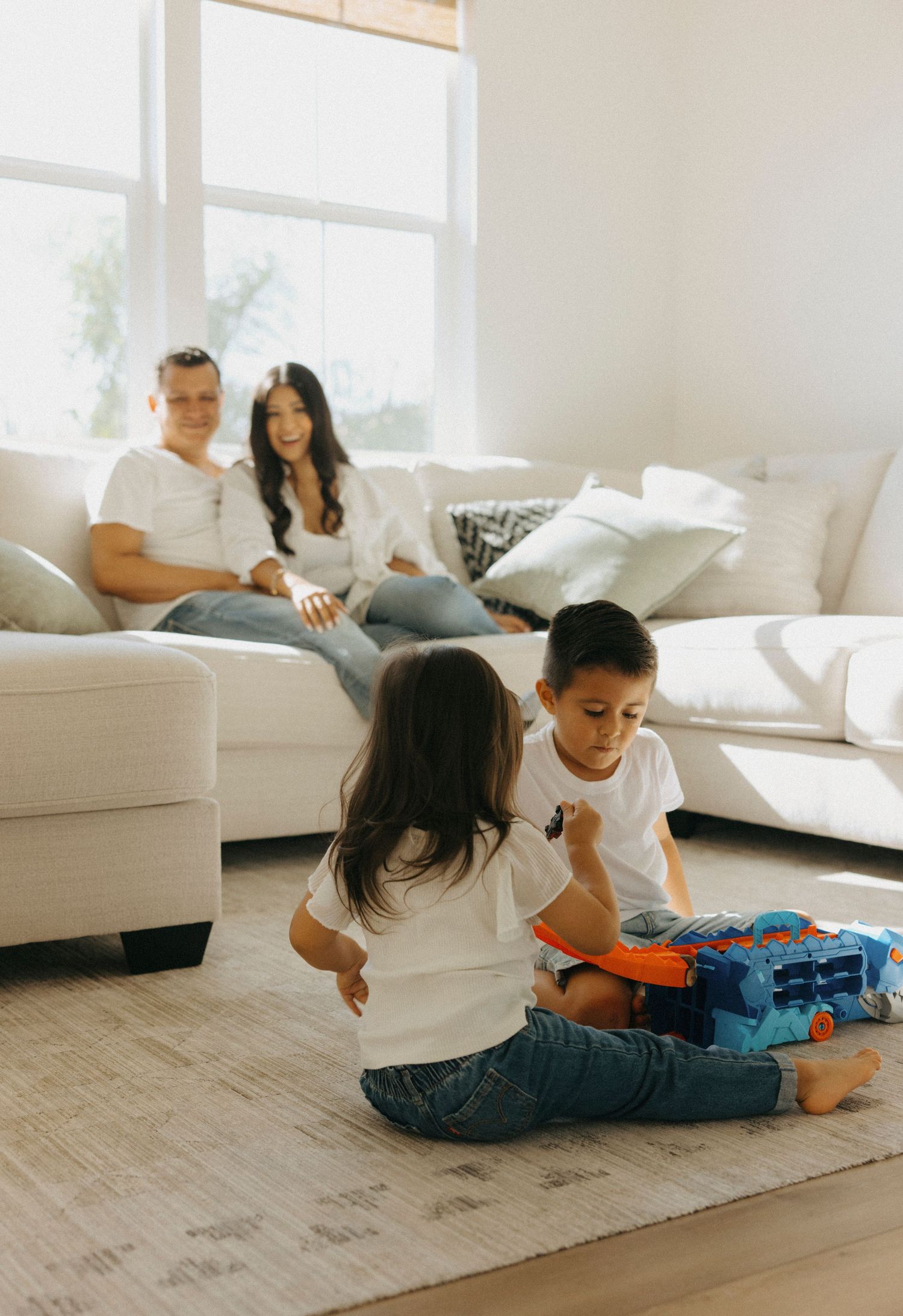 Family playing and lounging together on a light gray rug in a bright living room.