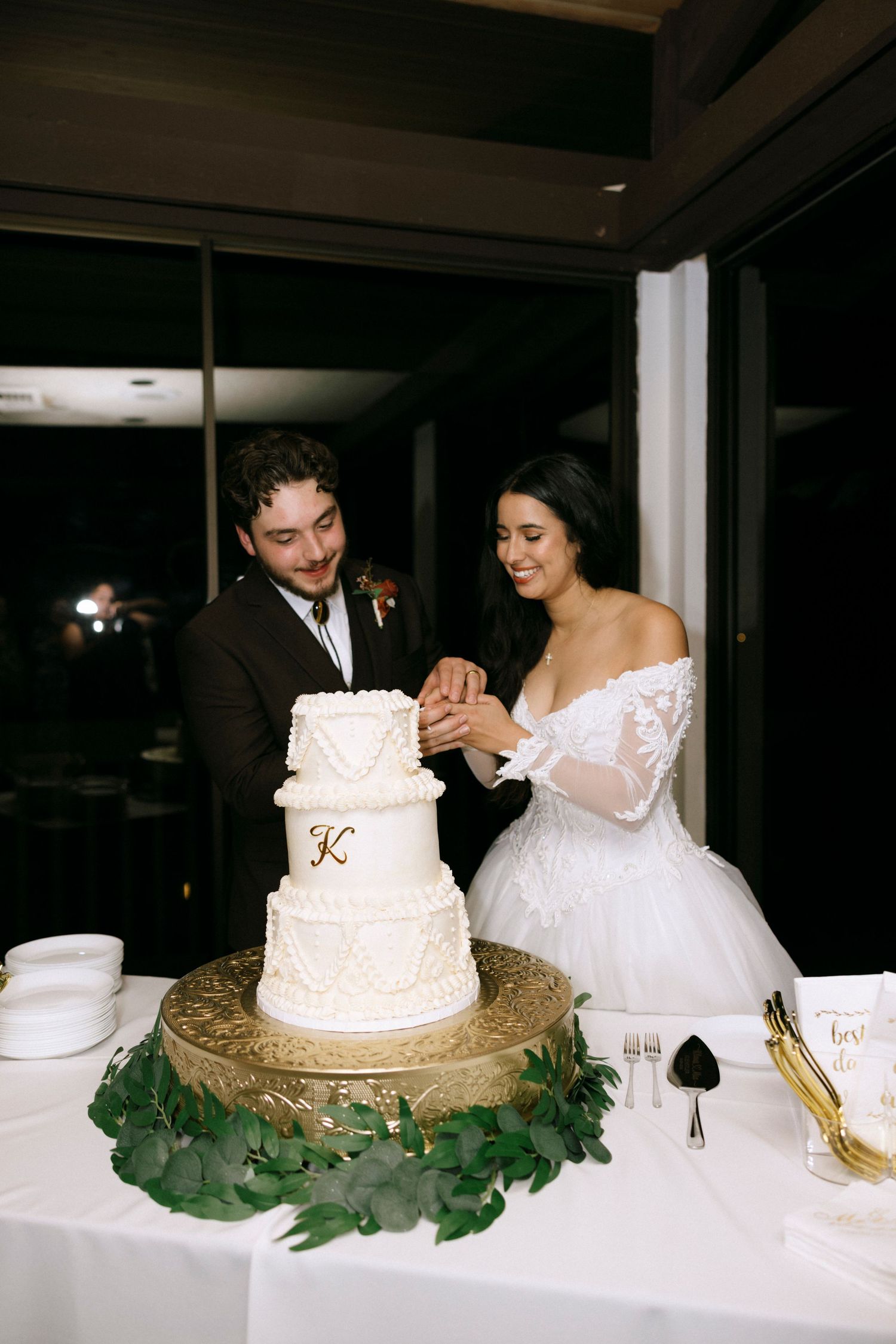 White wedding cake displayed on a gold stand with greenery decorations.