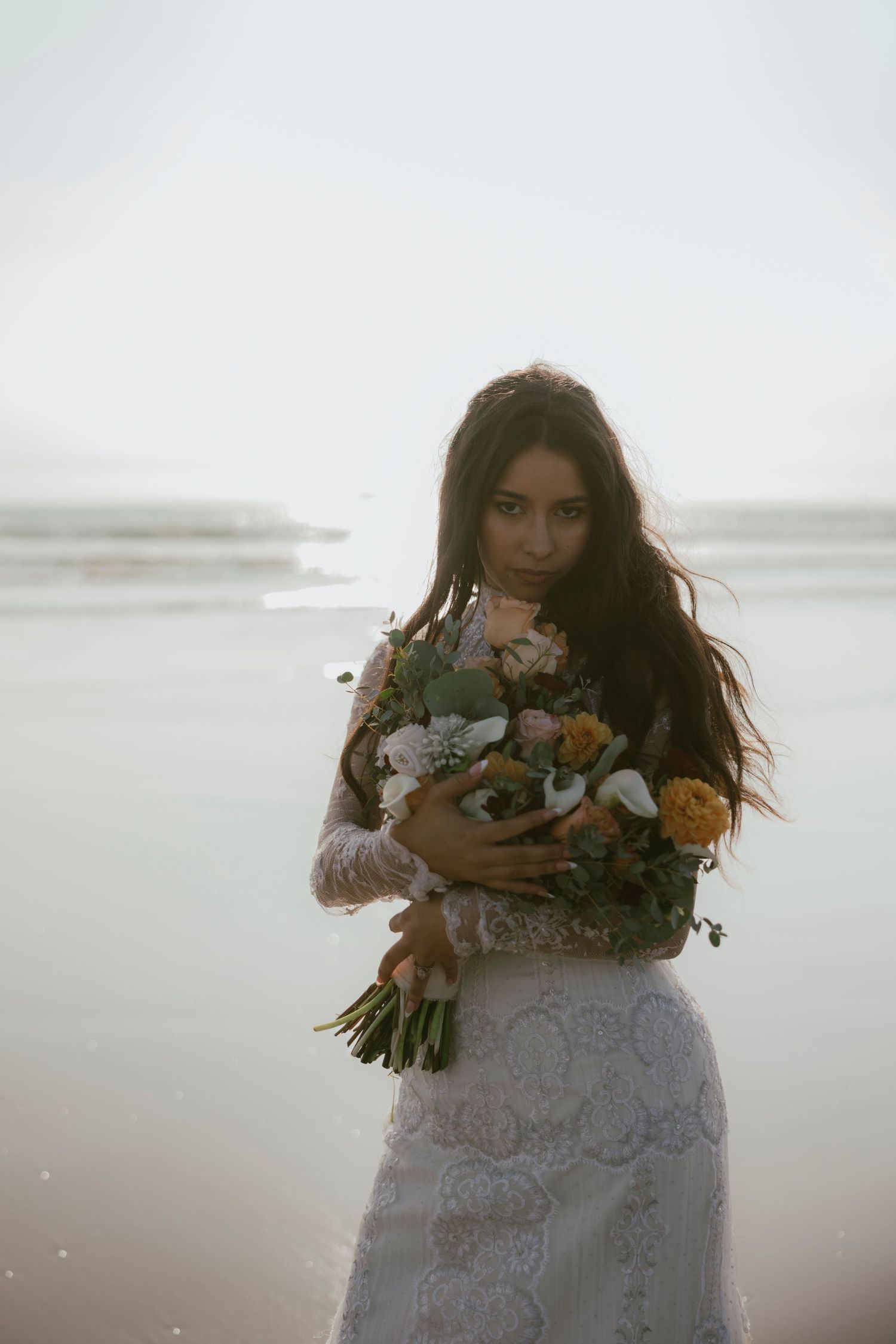 Bride in white lace gown holding bouquet on misty beach at sunset.