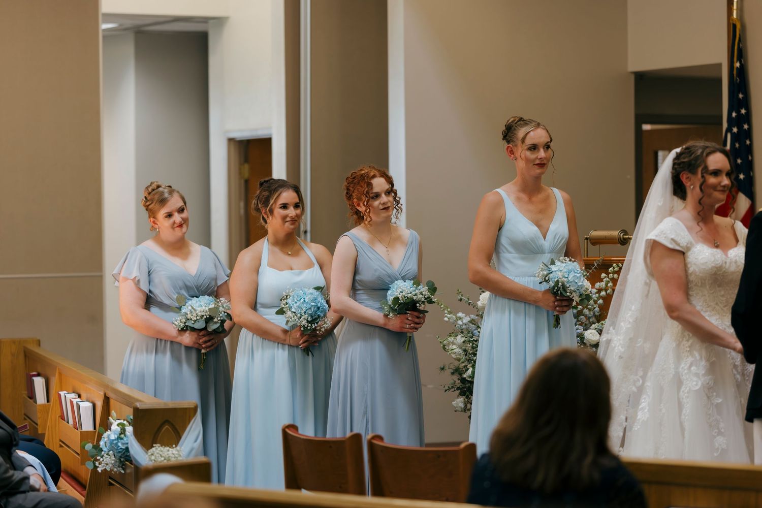 Four bridesmaids in light blue dresses holding white bouquets stand beside a bride in a white gown during a church wedding ceremony.