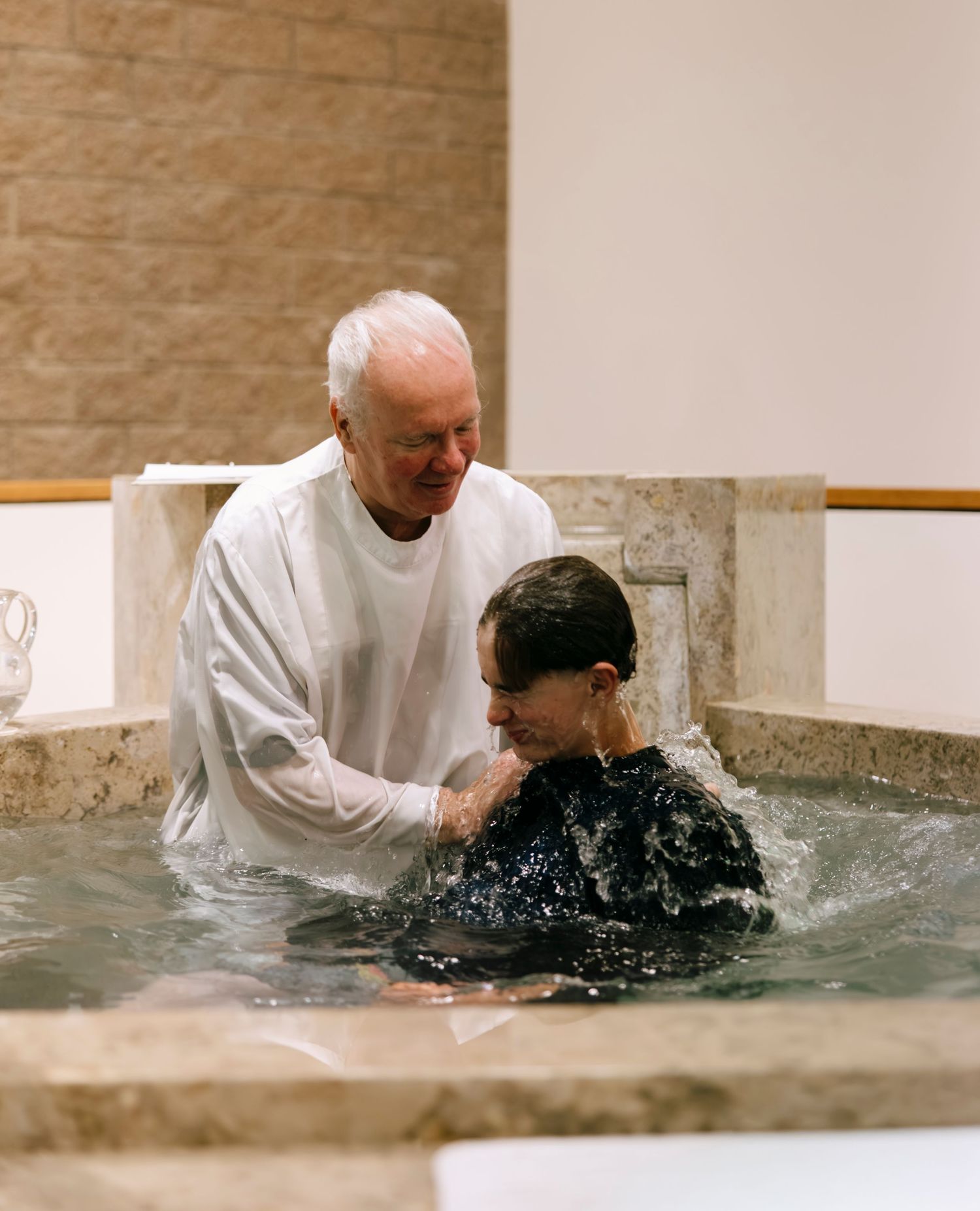 Priest performing full immersion baptism on adult candidate in baptismal font at church.
