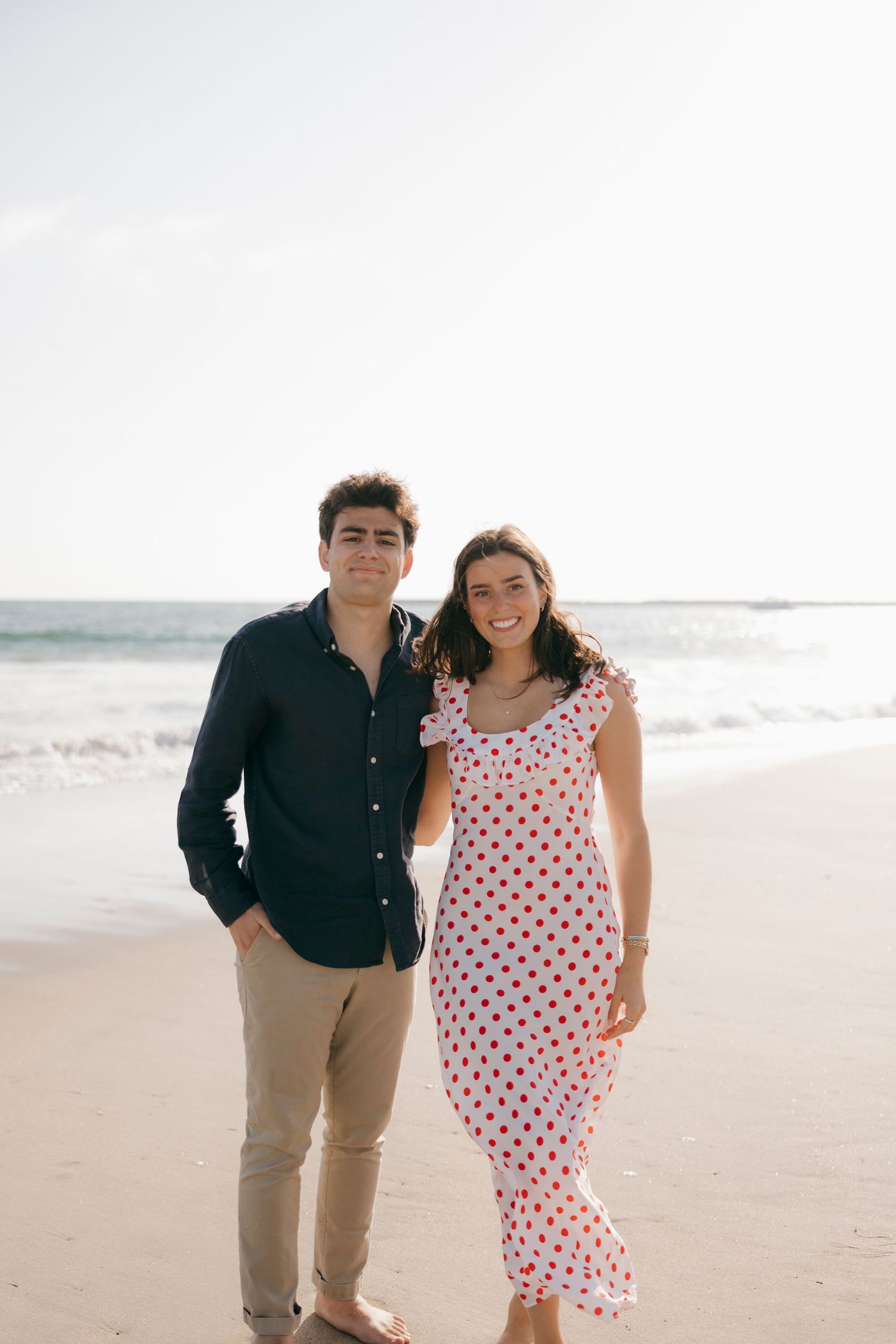 A smiling couple poses on a sandy beach, man in dark shirt and khakis, woman in white polka dot dress.